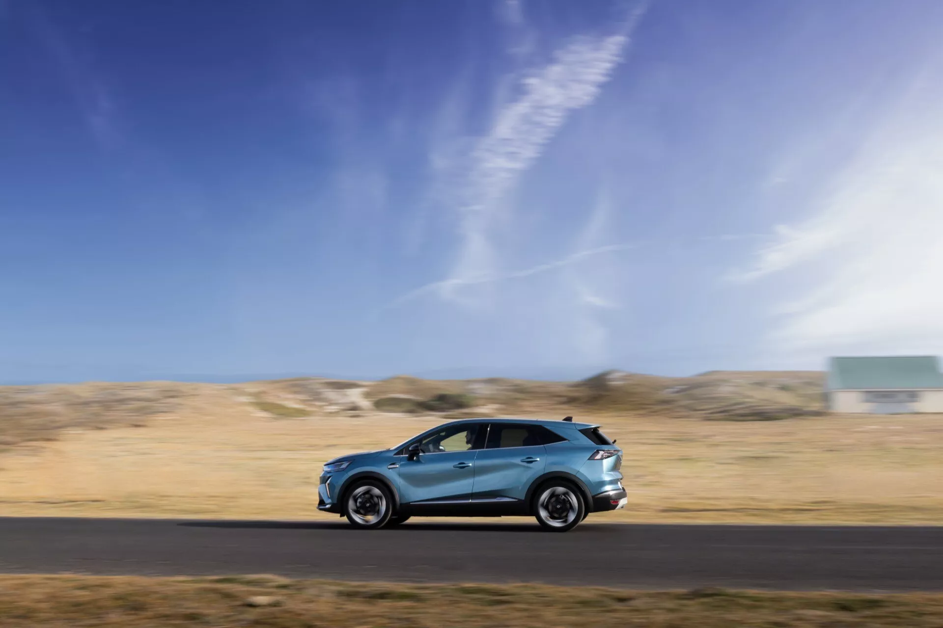 Dynamická bočná profilová fotografia pohybujúceho sa Renault Symbioz 2024 1. generácie, zachytená technikou panning na rozmazanom pozadí, ukazujúca jeho elegantný aerodynamický dizajn, zliatinové kolesá, bočné zrkadlá, integrované kľučky dverí a vysokokvalitný lak. Vozidlo je umiestnené v dolnej tretine rámu proti modrej oblohe a pustej krajine, čo zdôrazňuje rýchlosť, pohyb a schopnosť auta v rôznorodých prostrediach.
