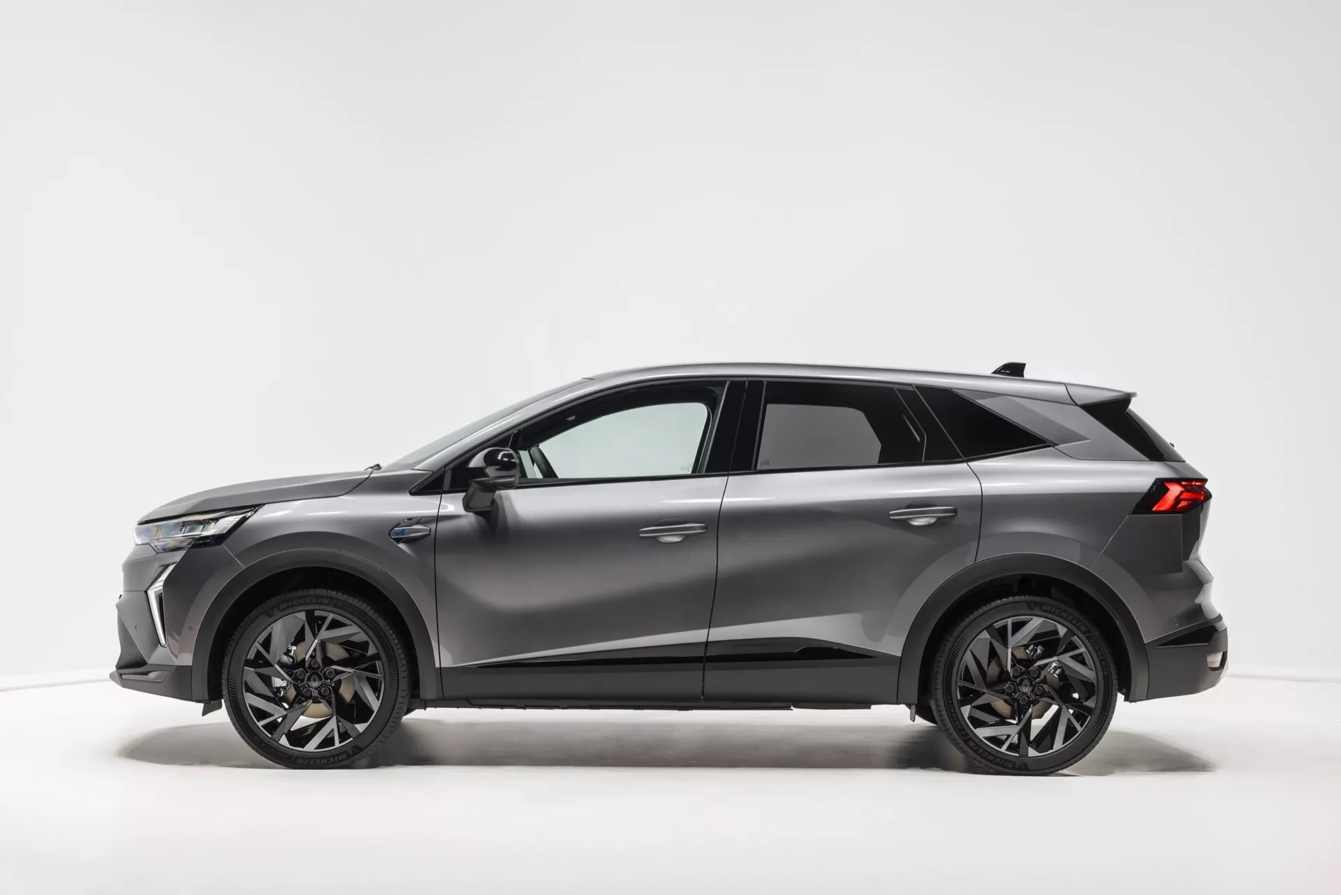 Obrázok ukazuje moderné SUV Renault Symbioz 2024 v štúdiovom prostredí s jednoduchým, svetlým pozadím zvýrazňujúcim vozidlo. Bočný profil zvýrazňuje dizajn auta s neutrálnym, kovovým sivým povrchom. Elegantná aerodynamika sa prejavuje strmo skoseným zadným oknom vedúcim k spoileru. Pätidverové SUV, navrhnuté na praktické použitie, sa môže pochváliť veľkými, tmavo zakončenými zliatinovými kolesami s viacramenným dizajnom, úzkymi LED svetlami, bočnými zrkadlami vo farbe karosérie s integrovanými smerovkami a strešnými nosičmi. Ďalšie podrobnosti zahŕňajú vyššiu svetlú výšku, anténu na zadnej časti strechy, mierne tónované okná a ochranné čierne obloženie na spodnej časti karosérie.