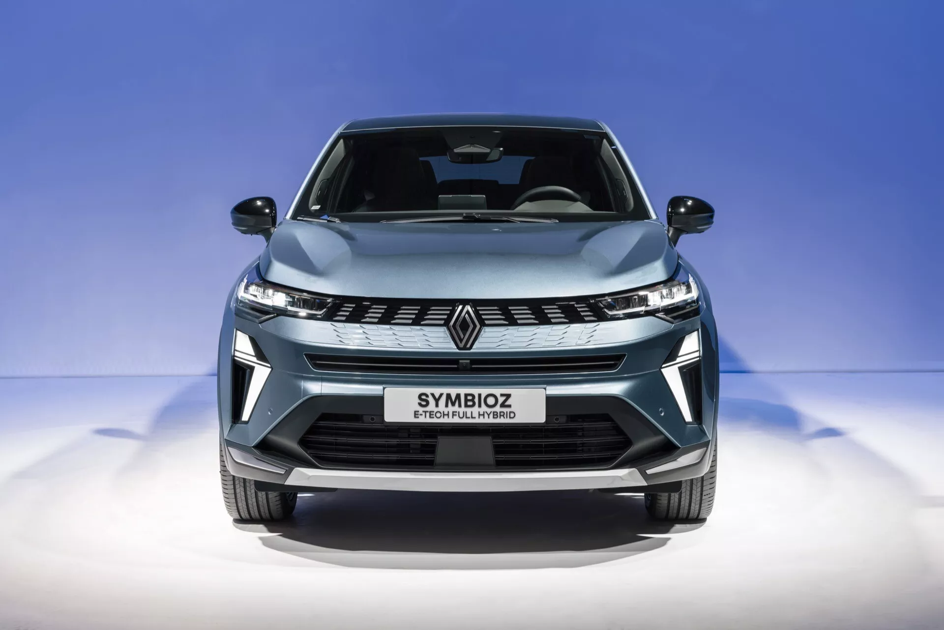 Blízky pohľad zepredu na Renault Symbioz 2024 (1. generácia) zachytený z mierne nízkeho uhla. Obrázok zdôrazňuje elegantnú prednú mriežku so štýlovým logom 'R', uhlové LED svetlomety a aerodynamicky tvarovanú kapotu. Metalická svetlomodrá farba auta a jeho lesklý povrch sú zvýraznené proti čistému, prechodnému modrému pozadiu. Text 'SYMBIOZ' na mieste pre evidenčné číslo naznačuje moderný a možno futuristický dizajn s prvkami ako LED osvetlenie, možné autonómne systémy a pokročilú konektivitu alebo elektrický pohonný systém.