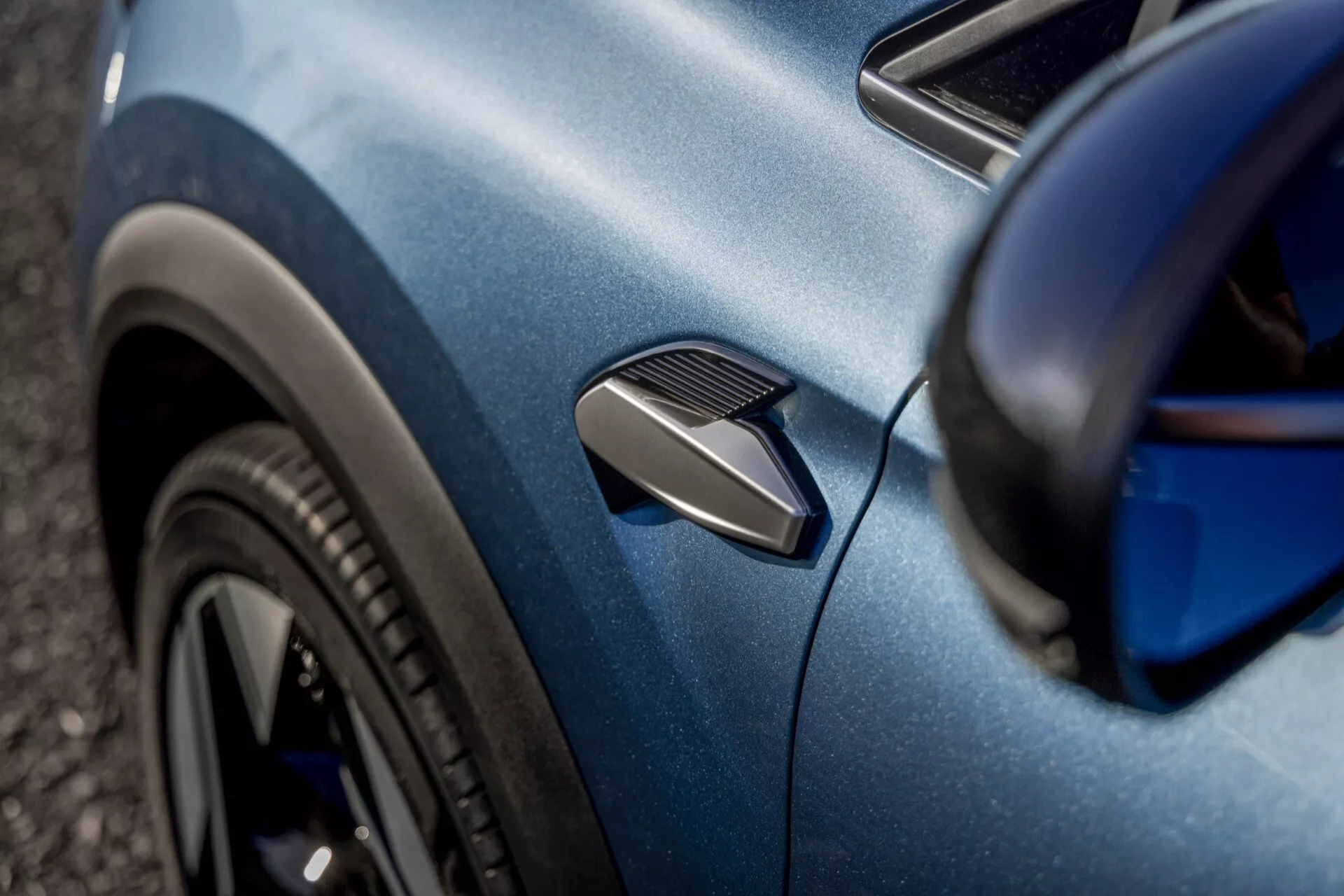 Detailný pohľad na modrý Renault Symbioz 2024 (1. generácia), zameraný na zakrivenie bočného panela. Fotografia zvýrazňuje funkčný bočný vetrací otvor, časť čierneho viacramenného predného kolesa a karosériu s kovovým povrchom. Plytký uhol a selektívne zaostrenie zdôrazňujú aerodynamické a dizajnové detaily auta s rozmazaným pozadím.