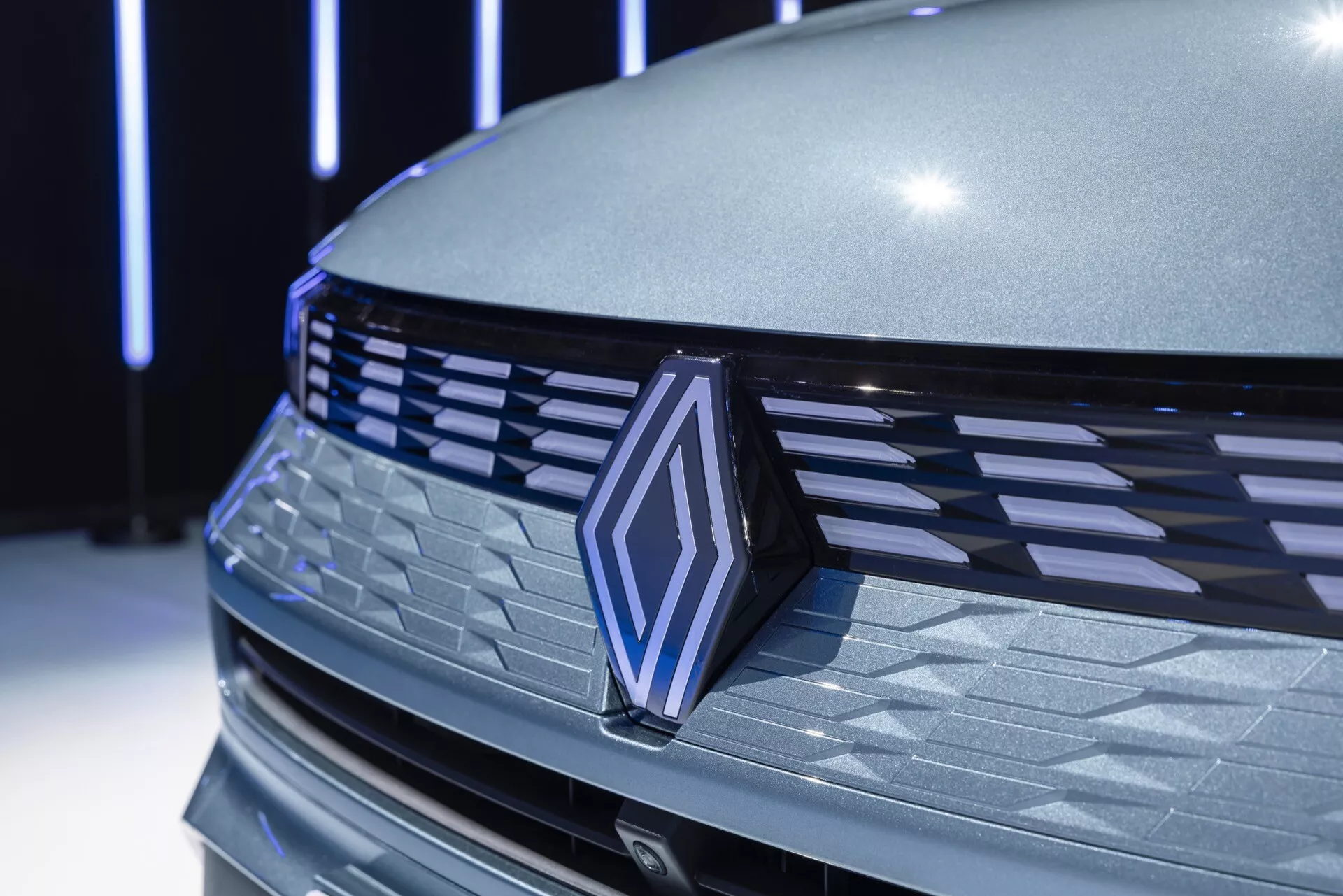 Detailný pohľad na prednú časť Renault Symbioz 2024, s dôrazom na mriežku s horizontálnymi lamelami, výrazný emblém s chrómovým okrajom a tvarovanú kapotu. Kovový šedý povrch odráža mäkké vnútorné osvetlenie, čím zdôrazňuje aerodynamický a luxusný vzhľad auta. Možné senzory pre asistenčné systémy vodiča môžu byť skryté v symetrickej mriežke, čo naznačuje pokročilú technológiu.