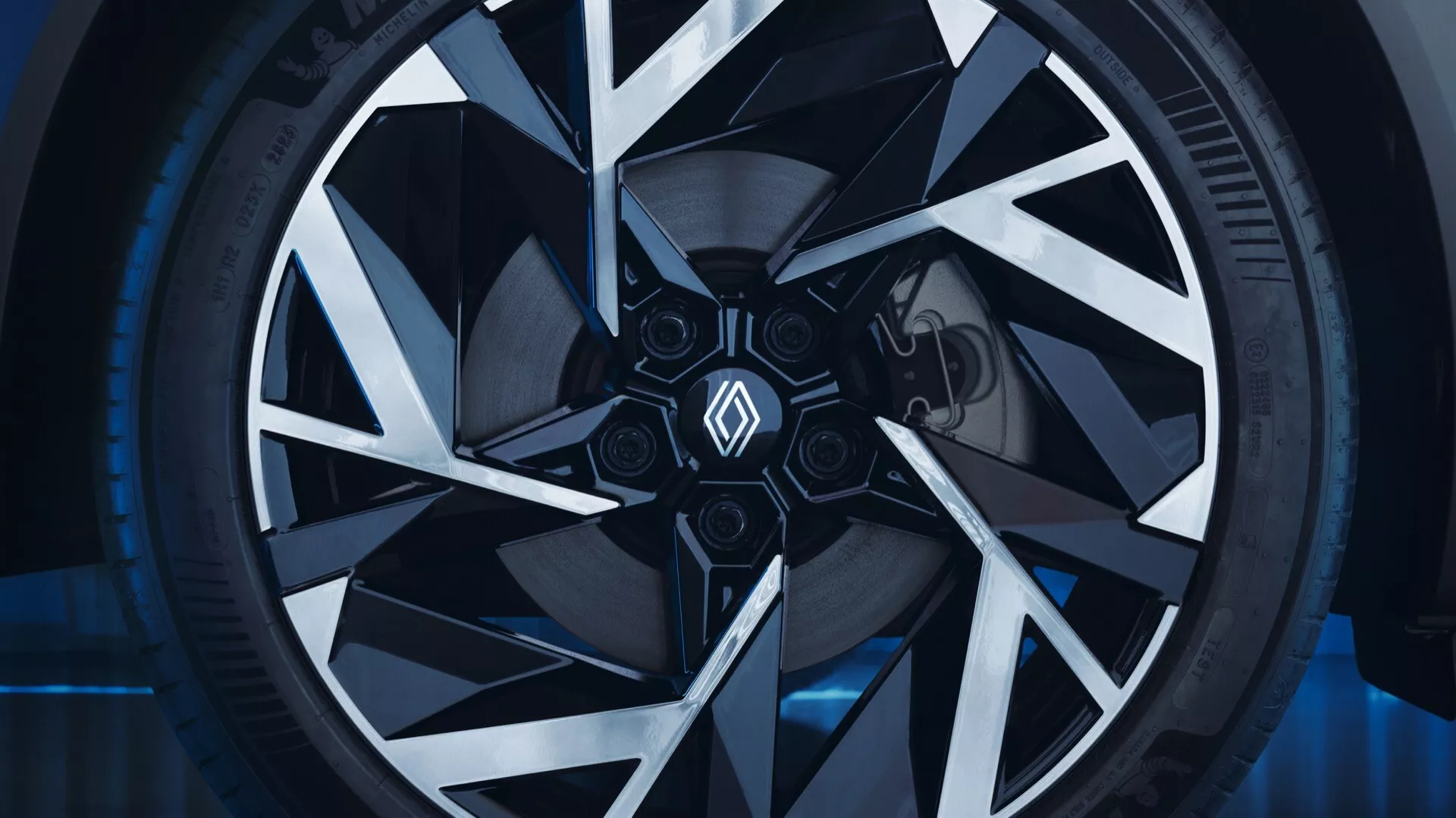Zbližovací bočný pohľad na koleso Renault Symbioz 2024 prvej generácie, zdôrazňujúci jeho zložitý geometrický dizajn ráfiku so striebornými a čiernymi uhlovými vzormi hviezd a nízkoprofilovú pneumatiku v kontrolovanom prostredí showroomu s modrým okolným osvetlením.