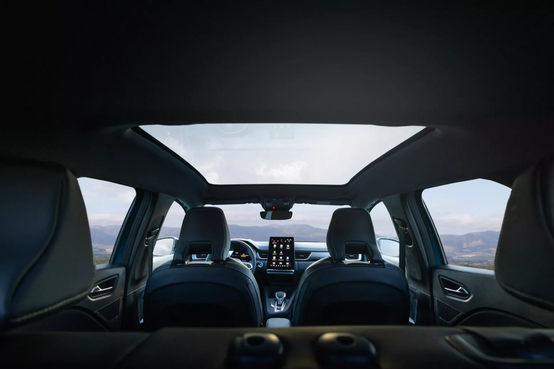 Záber interiéru vozidla Renault Symbioz 2024 (1. generácia) z pohľadu zadného sedadla, ktorý zobrazuje moderný dizajn s koženým čalúnením, výrazným šitím a pohodlne tvarovanými prednými sedadlami s opierkami hlavy. Na stredovej palubnej doske sa nachádza výrazný vertikálny dotykový infotainment systém, ovládacie gombíky klimatizácie a digitálny displej alebo HUD na vrchu. Multifunkčný volant je čiastočne viditeľný. Pohľad cez čelné sklo ukazuje horskú krajinu pod zamračenou oblohou, čo naznačuje, že vozidlo stojí na malebnom mieste.