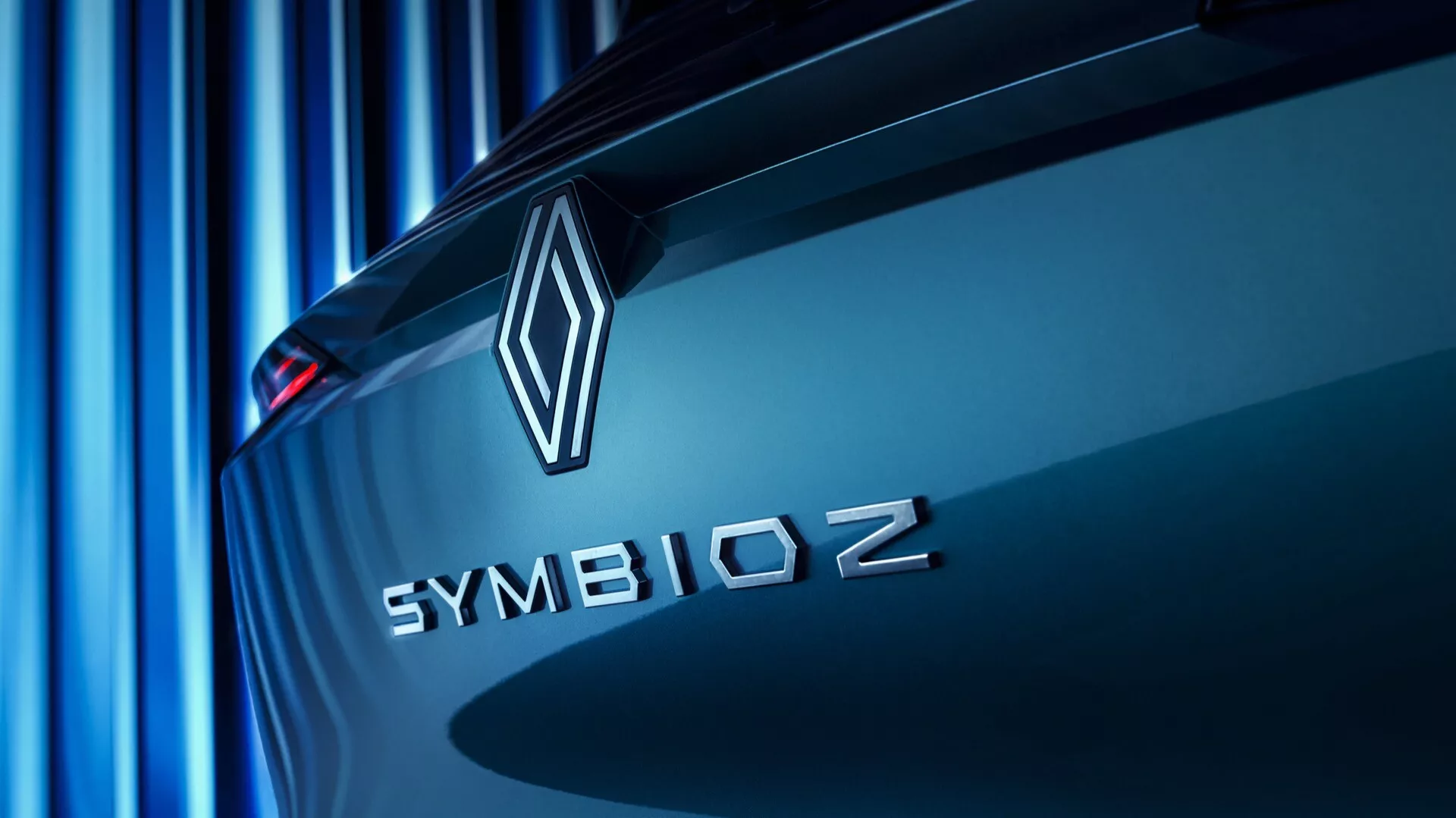 Zblízka a pod uhlom zobrazený zadný pohľad na vozidlo Renault Symbioz 2024 (1. generácia), so zameraním na značkový emblém a názov modelu. Emblém v tvare diamantu a 'SYMBIOZ' veľkými písmenami bezpatkovým písmom sú jasne viditeľné na poklope kufra, čo naznačuje dôraz na inovácie a technológie. Jemný modrý odtieň vrhá paralelné línie v pozadí, čím vytvára modernú a elegantnú atmosféru. Vozidlo sa zdá byť nehybné, zvýrazňujúc svoje aerodynamické dizajnérske línie.