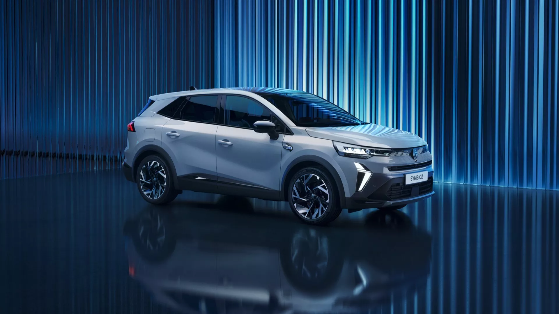 Digitálny štúdiový obrázok 2024 Renault Symbioz 1. generácia, moderného SUV s elegantným a dynamickým dizajnom. Profilový pohľad z mierneho predného uhla ukazuje bočný profil, prednú časť, mriežku, svetlomety, kolesá a bočné zrkadlá. Centrálne umiestnenie v ráme, fotografia z nízkeho uhla zdôrazňuje silný postoj auta. Ostré, uhlové svetlomety naznačujúce LED technológiu a denné svetlá. Úzka vodorovná mriežka zdôrazňujúca aerodynamiku. Tvarovaná charakterová línia tiahnúca sa od predného krídla po zadnú časť, ktorá prispieva k aerodynamickému vzhľadu. Viaclúčové zliatinové kolesá pridávajú na športovom vzhľade. Reflexný povrch pod autom a modré abstraktné digitálne pozadie vytvárajú prémiovú, futuristickú estetiku, možno naznačujúcu modernú alebo dokonca elektrickú pohonnú technológiu.