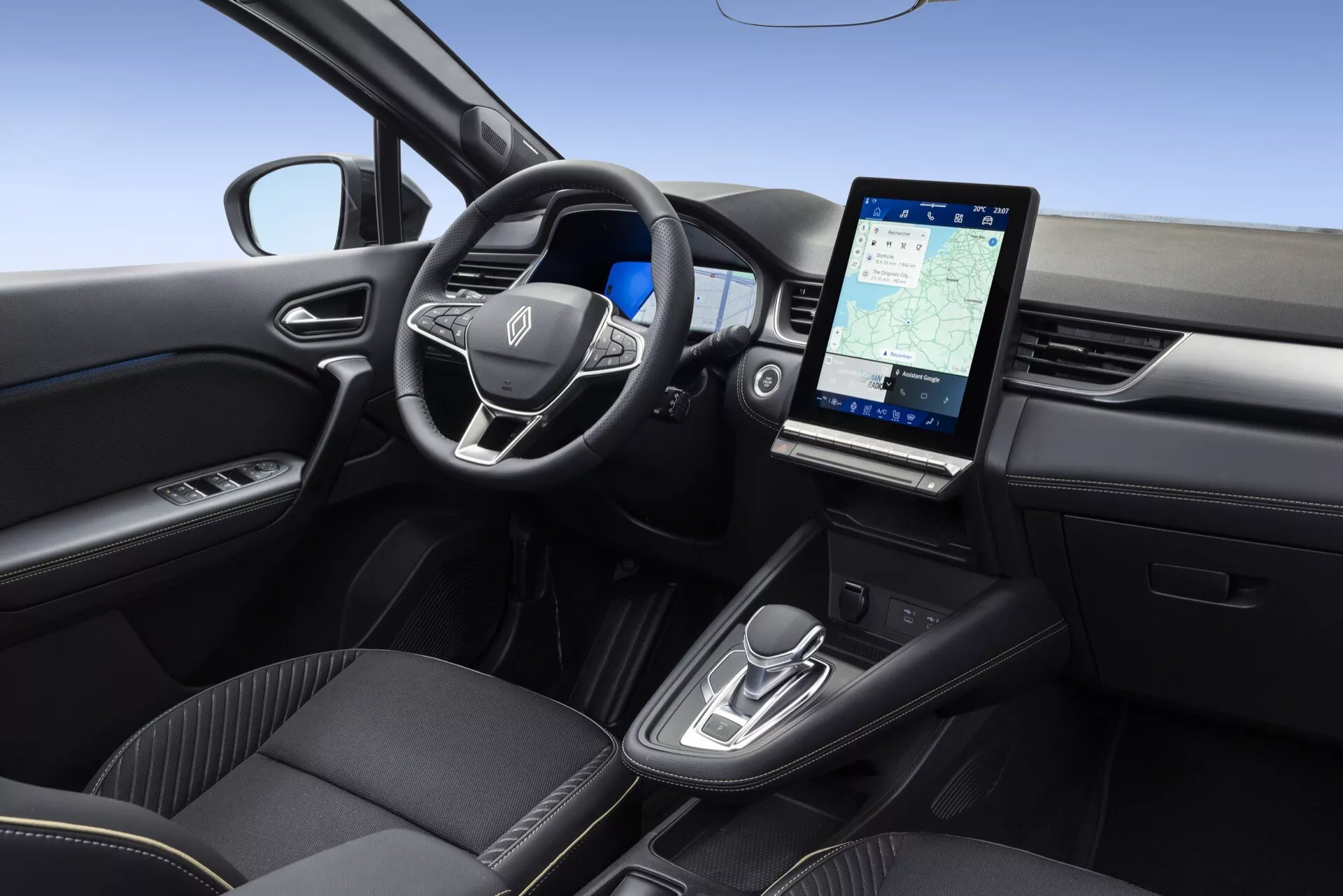 Interiér modelu Renault Symbioz 2024, zachytený z vyvýšeného, mierne pravého uhla. Obsahuje multifunkčný volant s emblémom výrobcu, minimalistický prístrojový panel naznačujúci digitálny alebo head-up displej, veľký vertikálny dotykový infotainment systém zobrazujúci navigáciu, moderné ovládanie automatickej prevodovky, vysoko kvalitné čierne sedadlá s prémiovým čalúnením a dvere vodiča s ovládaním elektrických okien.