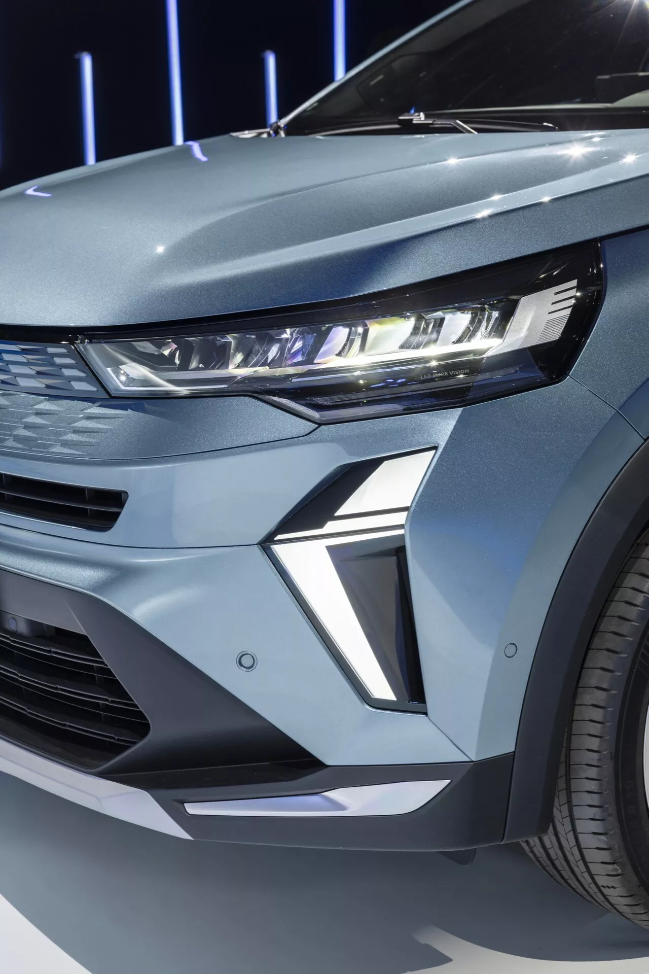 Detailný pohľad na prednú ľavú štvrtinu Renault Symbioz 2024 prvej generácie. Obrázok zachytáva moderný LED svetlomet vozidla s osvetleným denným svietením (DRL). Úzke teleso svetlometu sa tiahne do blatníka, čo exemplárne dopĺňa súčasný automobilový dizajn. Vysoce tvarovaný spodný diel pod svetlometom pridáva vozidlu futuristický a agresívny vzhľad. Viditeľný segment prednej mriežky naznačuje horizontálne nasávanie alebo dekoratívny panel, ktorý zapadá do športovej estetiky. Kovový povrch vozidla a viditeľné viacramenné koleso posilňujú jeho prémiový dojem.