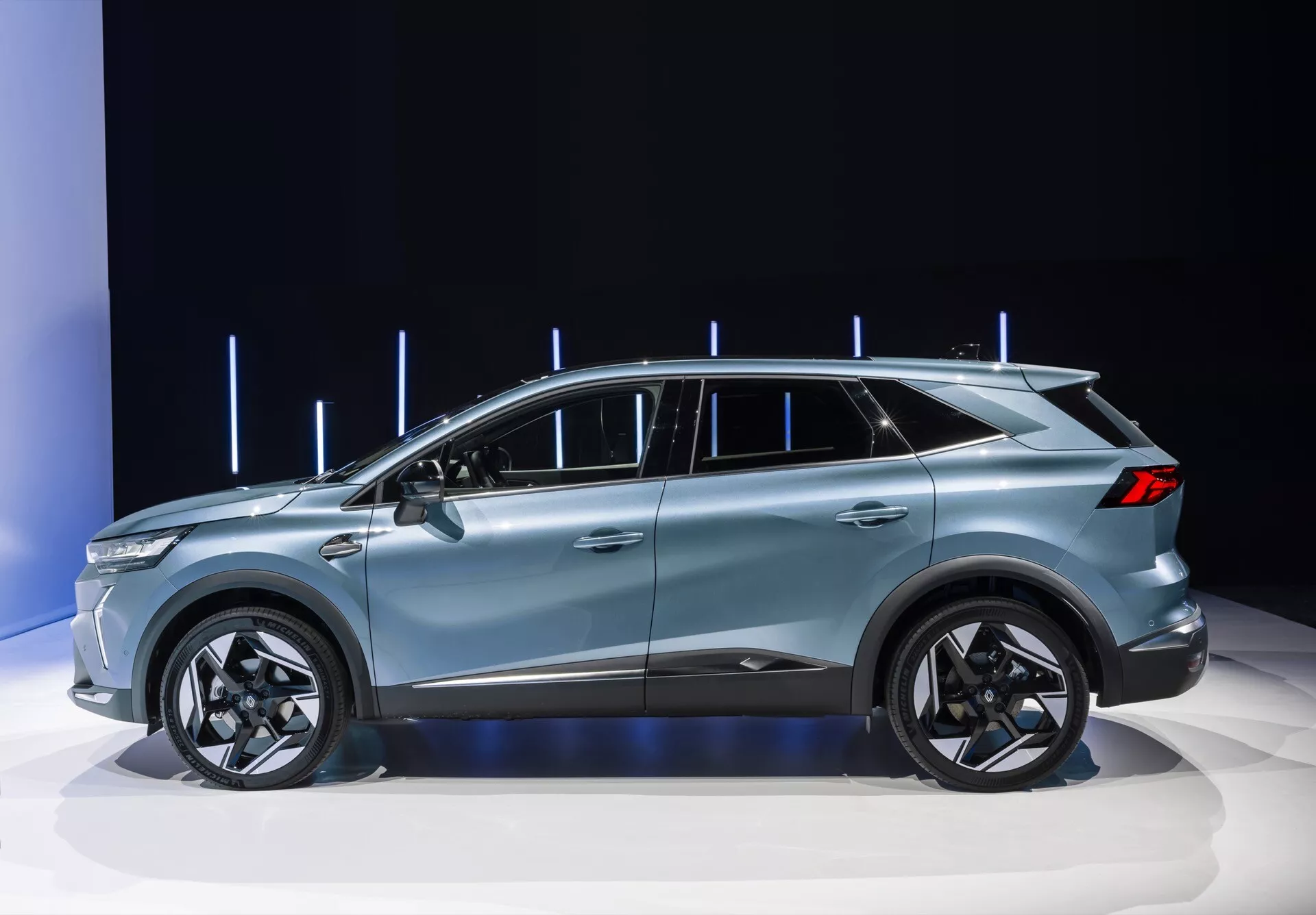 Obrázok zobrazuje Renault Symbioz 2024 (1. generácia) na výstavisku automobilov, výrazne zobrazujúc elegantný bočný profil moderného crossover SUV. Vozidlo sa vyznačuje kupé podobnou strechou, konfigurovaním s piatimi dverami, veľkými hliníkovými kolesami a sofistikovanými dizajnovými líniami, všetko zvýraznené čistým, dobre osvetleným pozadím so zvislými svetelnými prvkami. Dizajn naznačuje prémiové funkcie, ako sú LED technológia, aerodynamická účinnosť a prípadne pokročilé asistenčné technológie pre šoféra.