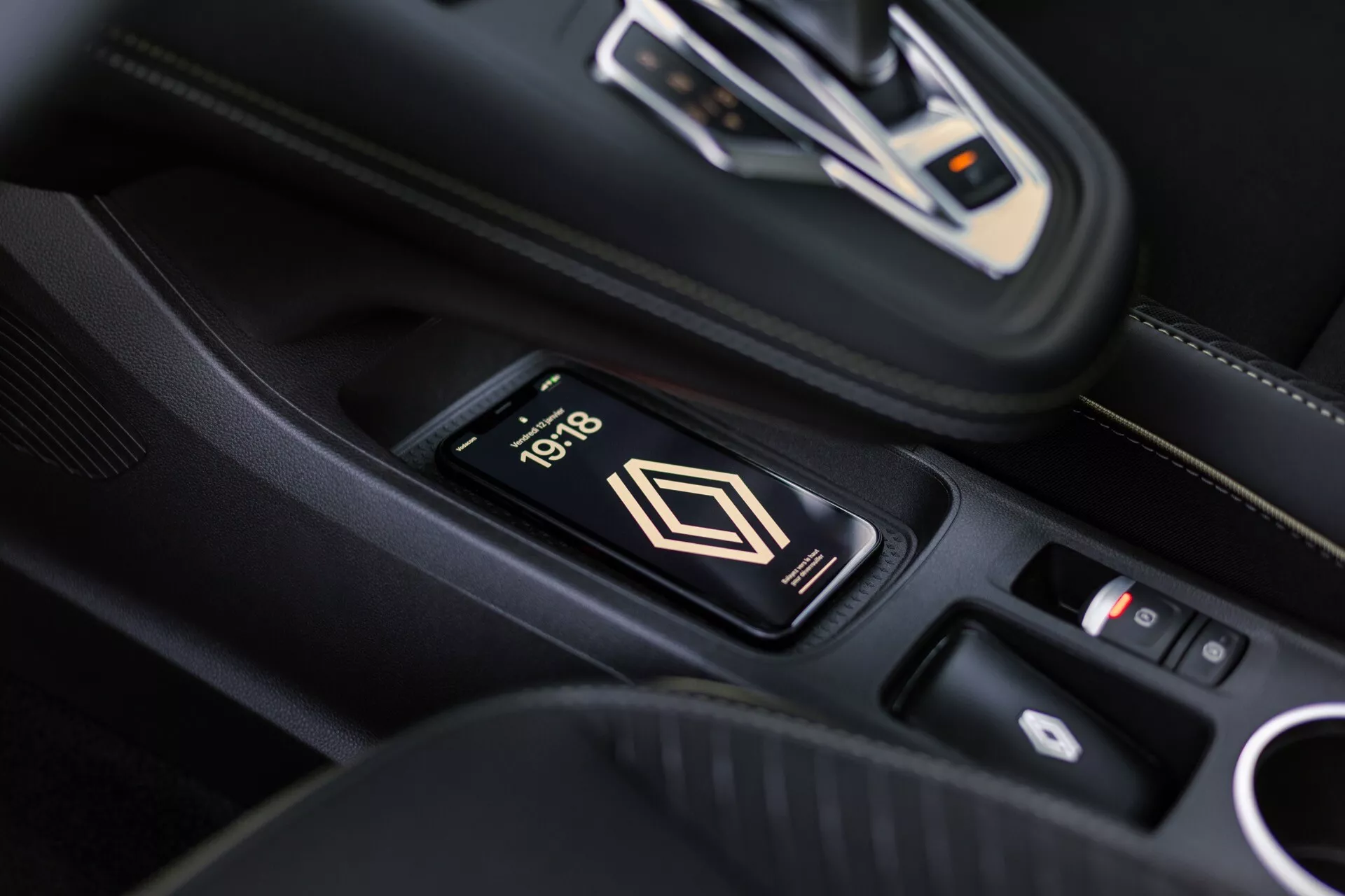 Pohľad do interiéru Renault Symbioz 2024 na stredovú konzolu s bezdrôtovou nabíjačkou, na ktorej je telefón ukazujúci čas 10:18. Elegantná automatická prevodovka s kovovými akcentmi a ovládací panel v pozadí. Prémiový interiér s čiernou kožou a bielym prešívaním.