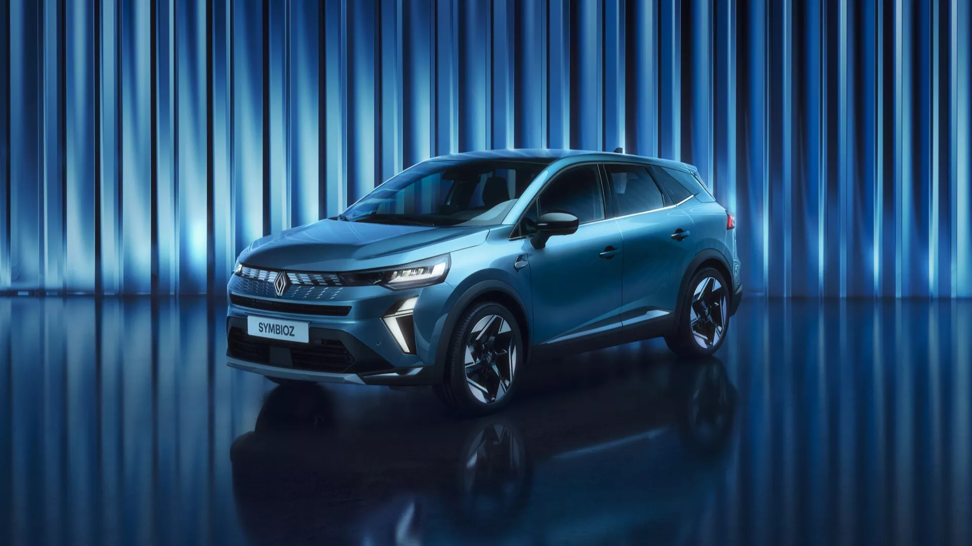 Obráz zobrazuje SUV Renault Symbioz 2024 (1. generácia) umiestnené v štúdiu s dynamickým modrým pozadím s vertikálnymi svetelnými pruhmi. Snímka z nízkeho až stredného uhla zachytáva impozantný predok a elegantný bočný profil vozidla. Celé vozidlo je na zábere, zdôrazňujúc jeho prítomnosť a moderný dizajn. Viditeľné sú ostré, hranaté LED svetlomety a výrazná mriežka, spolu s hladkým predným nárazníkom, ktorý pravdepodobne obsahuje bezpečnostné senzory. Bočný profil odhaľuje aerodynamické línie, zapustené kľučky dverí, aerodynamické zrkadlá a veľké zliatinové kolesá. Kovový lak a športový postoj automobilu sú zvýraznené štúdiovým osvetlením a odrážajúcim povrchom pod ním.