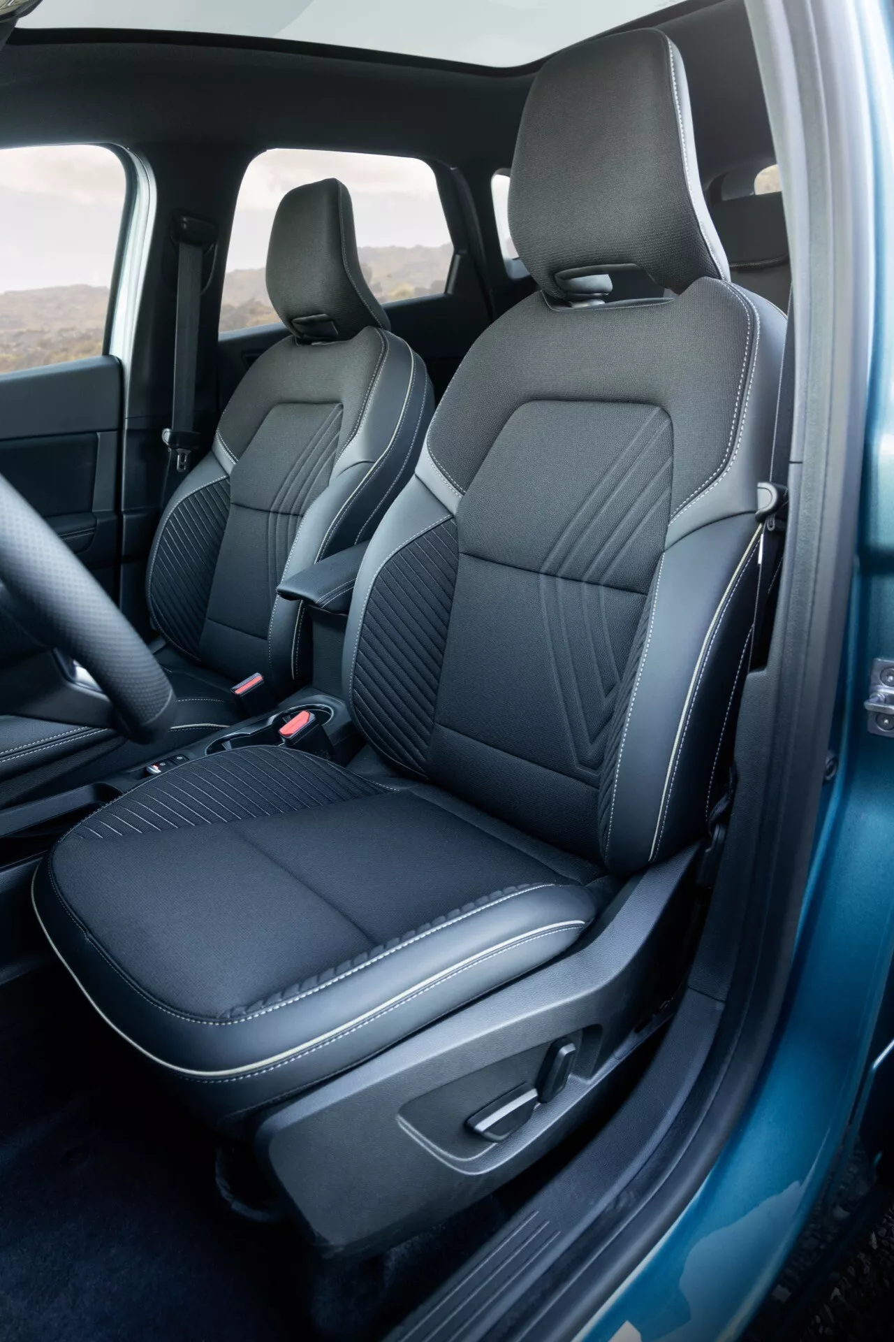 Detailný záber na interiér modelu Renault Symbioz 2024 prvej generácie, zameraný na predné sedadlo spolujazdca a časť sedadla vodiča. Fotografia je urobená z šikmého uhla, ktorý zvýhodňuje stranu spolujazdca a ukazuje tmavé, prémiové čalúnenie s detailným vzorom štepovania. Otvorené dvere spolujazdca odhaľujú ergonomický dizajn sedadiel, hladké bočné opierky a centrálne oblasti s diamantovým prešívaním, ktoré zdôrazňujú kombináciu luxusu a športového charakteru. Integrované opierky hlavy a minimalistický dizajn bez viditeľných nastavovacích tlačidiel nasvedčujú čistému, modernému vzhľadu. Panel otvorených dverí zodpovedá materiálu sedadla, s lakťovou opierkou a rozmazanými ovládacími prvkami. Tmavý koberec doplňuje sedadlá. Viditeľný B-stĺpik a gumové tesnenia rámu dverí naznačujú pozornosť venovanú zvukovej izolácii, ktorá naznačuje vysokokvalitný dizajn.