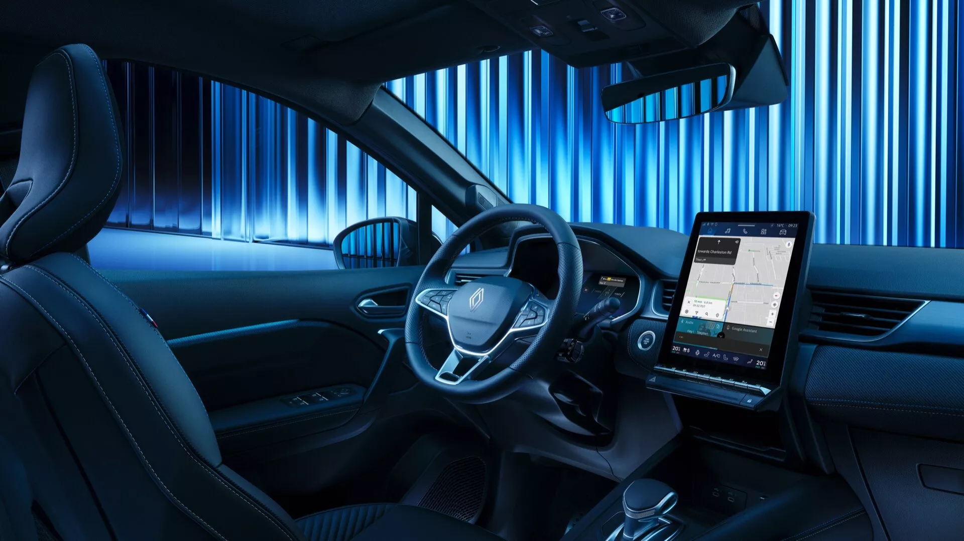 Pohľad zo zadného sedadla na interiér Renaultu Symbioz 2024, ktorý zvýrazňuje centrálne umiestnený volant s integrovanými ovládacími tlačidlami, digitálny prístrojový panel a prominentnú vertikálne orientovanú infotainment obrazovku zobrazujúcu mapu. Kabína má modré ambientné osvetlenie, ktoré naznačuje moderný a technologicky orientovaný dizajn, minimálne fyzické tlačidlá na stredovej konzole a časť dverového panelu s integrovaným reproduktorom, čo naznačuje prémiový zvukový systém.
