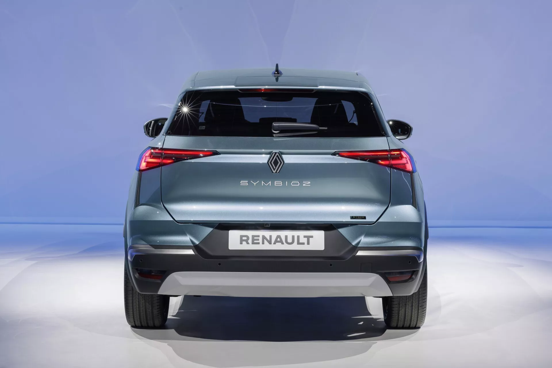 Zadný pohľad na Renault Symbioz 2024, ktorý ukazuje svoje moderné dizajnové prvky vrátane výrazných LED zadných svetiel, výrazného loga značky, názvu a modelu vyryté veľkými písmenami, zadného stierača, vysokomontovaného brzdového svetla a senzorov zadného nárazníka na modrom gradientovom pozadí.