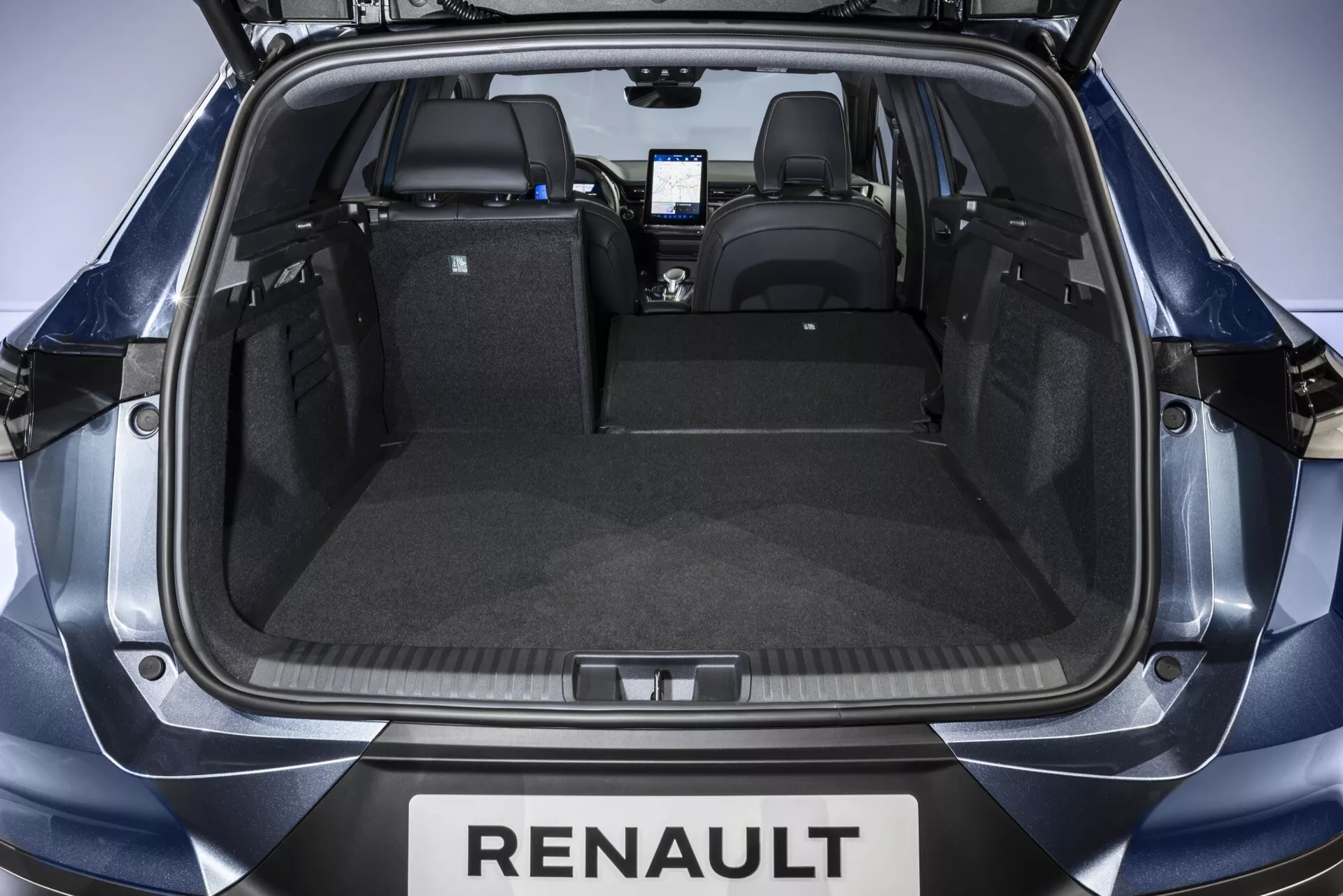 Pohľad do batožinového priestoru Renaultu Symbioz 2024 z vysokého uhla za otvoreným zadným vekom, ukazuje čistý, nezastavaný kufor so sedadlami v zvislej polohe, zadný nárazník s označením Renault, trvanlivé obloženie, zástrčku na 12 voltov na ľavej strane a úchyt na upevnenie predmetov na pravej strane. Viditeľný je interiér kabíny a obrazovka stredovej konzoly, s rovnomerným štúdiovým osvetlením zvýrazňujúcim prvky.