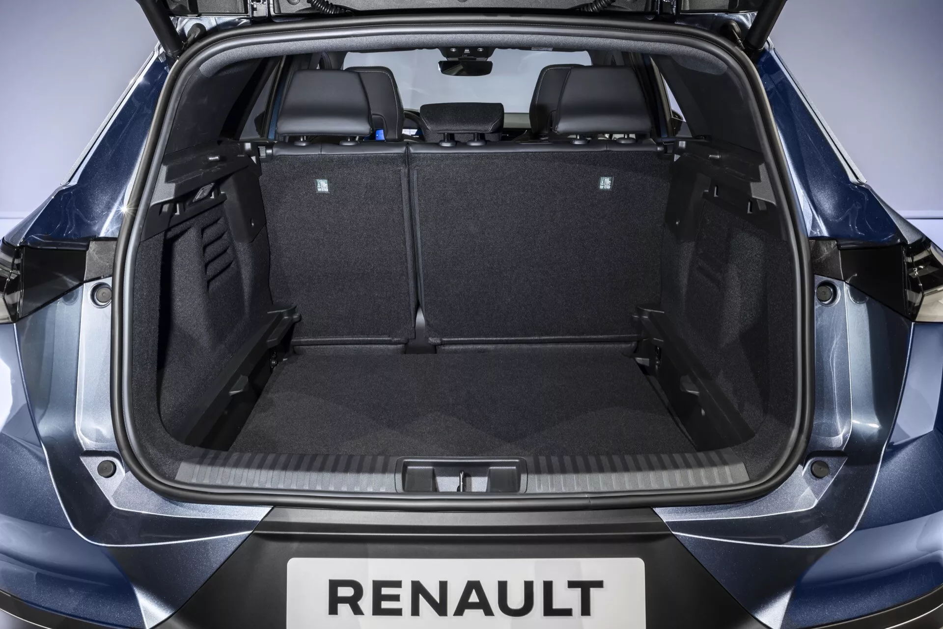 Vnútorný pohľad na úložný priestor Renault Symbioz 2024 z otvorenej zadnej brány, zobrazujúci symetrický priestor kufra s čiernym kobercom, plastovým obložením a sklopenými zadnými sedadlami na zväčšenie úložného priestoru. Viditeľný branding zahŕňa 'RENAULT' pod bránou.