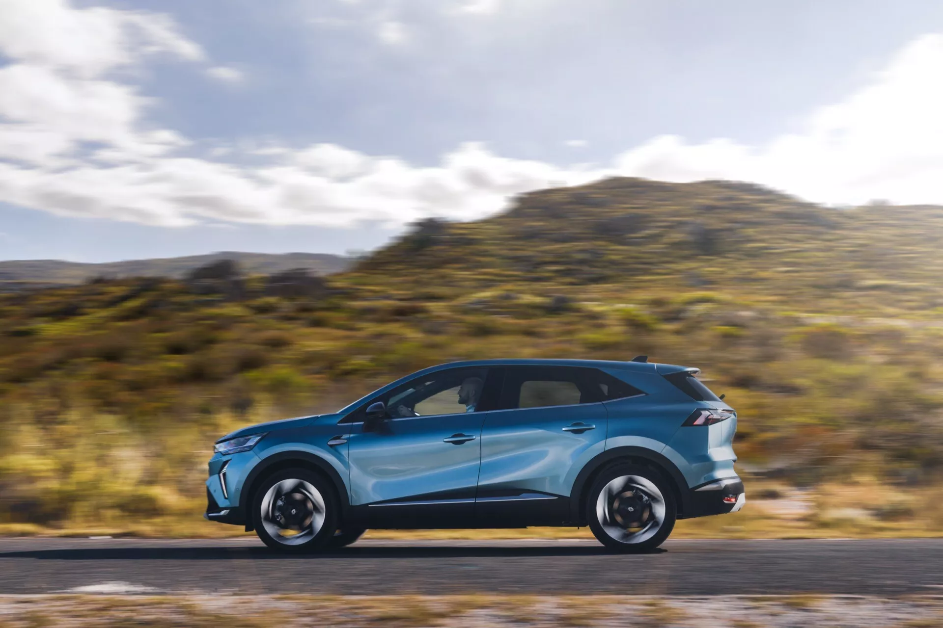 Svetlomodrý Renault Symbioz 2024 (1. generácia) v pohybe, ukazujúci svoje elegantné aerodynamické línie a moderný dizajn z profilu. Auto je umiestnené v ľavej tretine obrazu, má rozmazané pozadie a cestu, čo naznačuje rýchlosť. Viditeľné prvky zahŕňajú LED denné svietenie, integrované smerovky v spätných zrkadlách, panoramatickú strechu, výrazné ráfiky a nízkoprofilové pneumatiky. Fotografiu zachytená technikou panningu, ktorá zdôrazňuje dynamický pohyb auta.