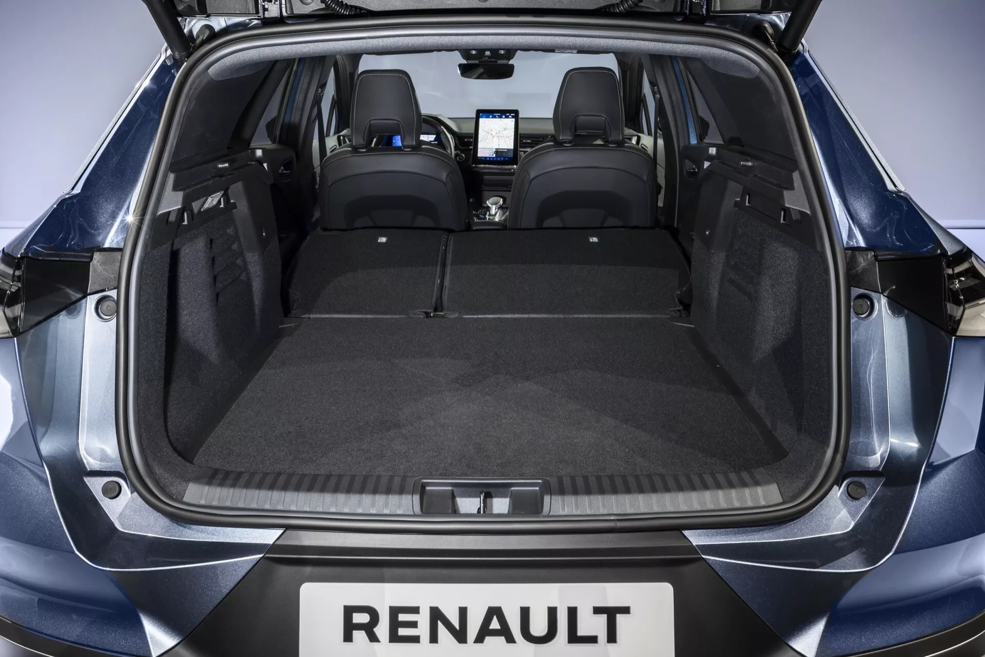 Pohľad zozadu otvorenými dverami do batožinového priestoru Renaultu Symbioz 2024. Priestranný kufor s plochou podlahou pokrytou šedým kobercom, navrhnutý bez prekážok pre maximálnu kapacitu. Dva zámky na oboch stranách naznačujú, že zadné sedadlá možno sklopiť pre viac miesta. Čierne kožené zadné sedadlá s opierkami hlavy a slabo viditeľný moderný infotainment systém v prednej kabíne naznačujú interiér vybavený technológiami.