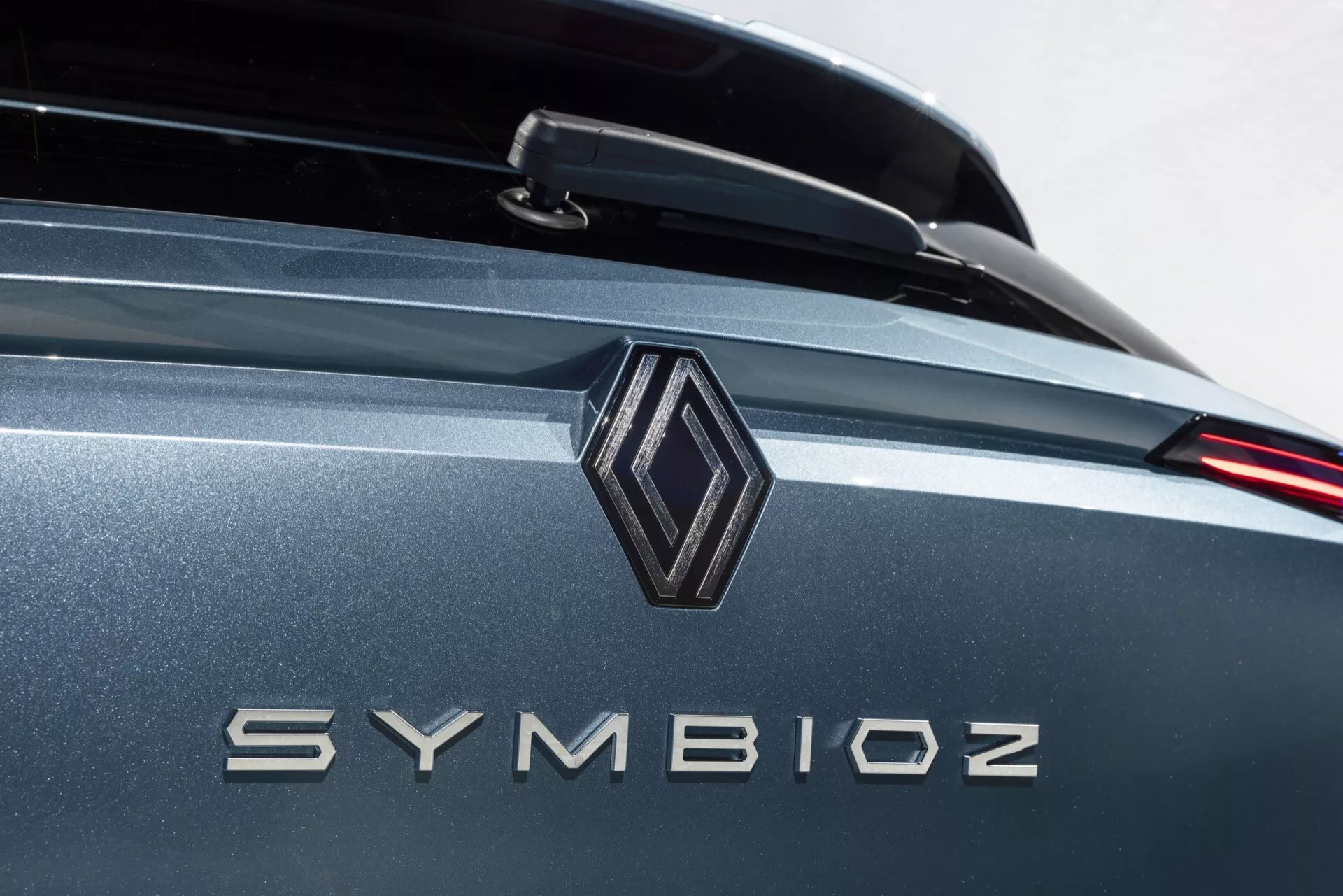 Profesionálny detailný záber zadnej časti auta zameraný na brandingové detaily s diamantovým emblémom a názvom modelu 'SYMBIOZ' v kovových písmenách. Obrázok využíva malú hĺbku ostrosti, čo zvýrazňuje emblém a nápis, zatiaľ čo pozadie jemne mizne. Dobre osvetlené prostredie naznačuje vnútorný showroom alebo štúdio. Farba vozidla je kovovo šedá alebo modrá s reflexnými vlastnosťami, obsahujúci možný zadný spojler a moderný dizajn LED koncových svetiel.