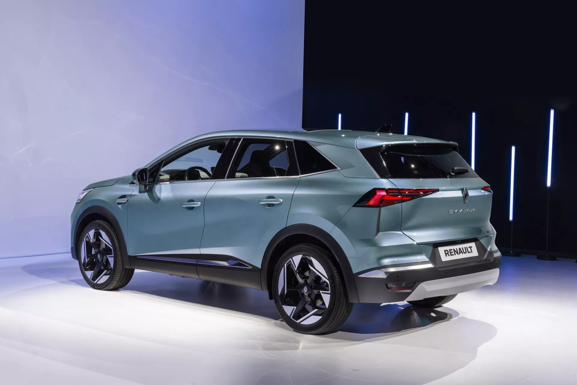 Trojštvrťový zadný pohľad na Renault Symbioz 2024. Tyrkysové SUV, umiestnené na výstavnej plošine, prezentuje moderný aerodynamický dizajn so zvažujúcou sa strechou, výraznými blatníkmi, ktoré obsahujú veľké zliatinové kolesá, a tónovanými zadnými oknami. Zreteľné LED zadné svetlá, zadný spojler, logo na zadnej strane a označenie modelu, parkovacie senzory a pravdepodobne zadnú kameru sú viditeľné. Elegantný zadný nárazník tiež obsahuje svetelné prvky, zatiaľ čo žraločia plutva naznačuje pokročilú technológiu. Integrované ukazovatele smeru vo vonkajších zrkadlách a zapustené kľučky dverí naznačujú bezkľúčový vstup. Minimalistické pozadie zvýrazňuje detaily vozidla.