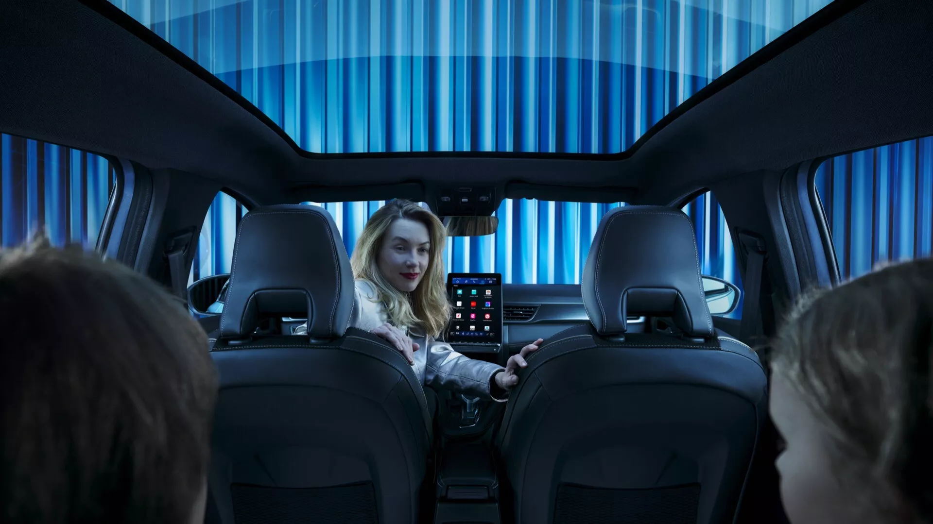 Pohľad na interiér Renault Symbioz 2024, prvej generácie, fotografovaný zo zadného sedadla smerom dopredu. Ukazuje zadnú časť dvoch predných sedadiel s nastaviteľnými opierkami hlavy, ženu na sedadle vodiča usmiatu smerom ku kamere a interaktívny displej umiestnený medzi sedadlami pre zadných cestujúcich. Rovnomerne osvetlený, studený modrý interiér naznačuje modernú, luxusnú atmosféru, ktorá je zvýraznená tónovanou panoramatickou strechou.