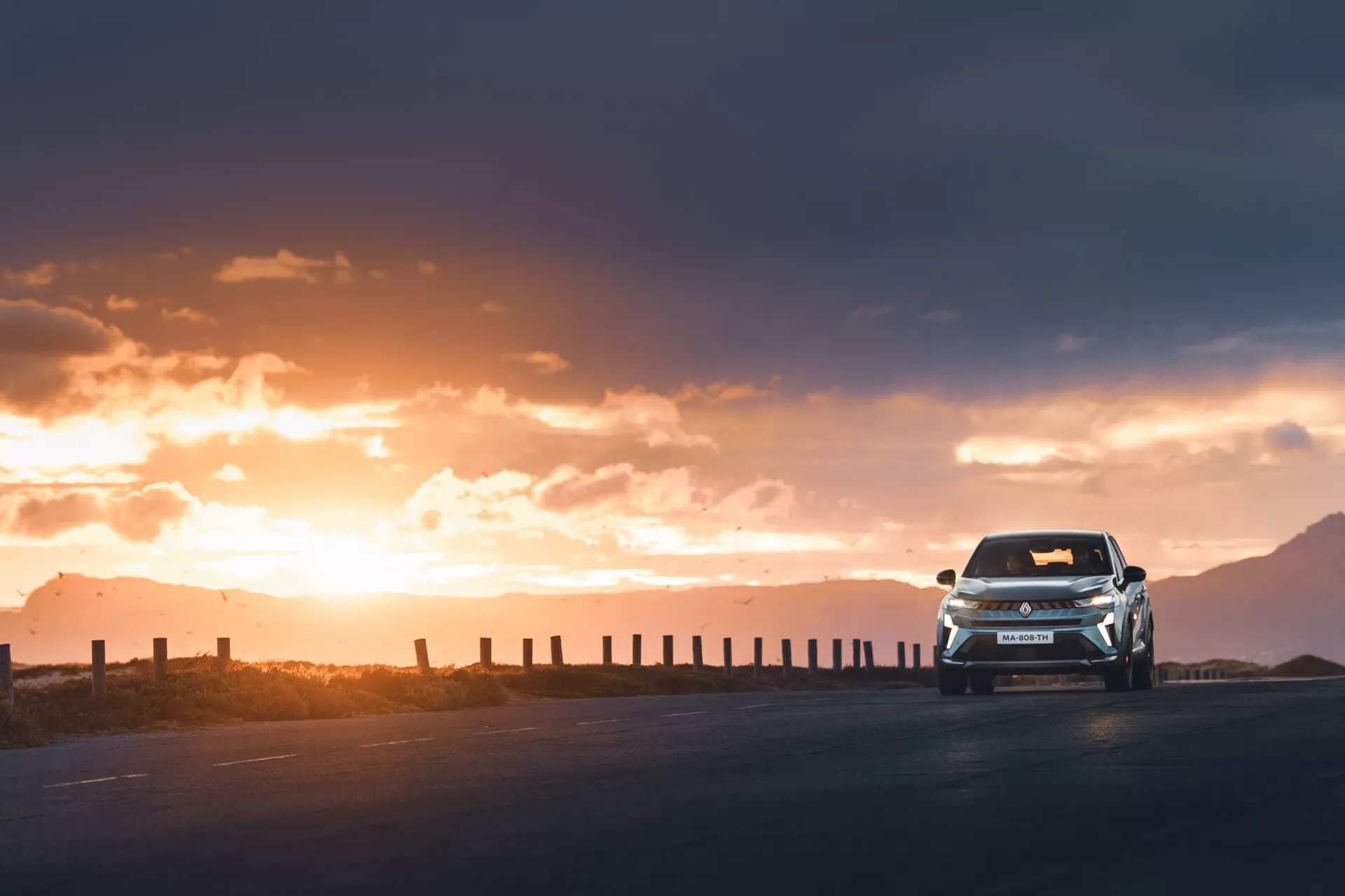 Obrázok zachytáva Renault Symbioz 2024 z nízkeho uhla, čo zdôrazňuje jeho dynamickú a silnú prítomnosť pri jazde smerom k fotoaparátu. Fotografia ukazuje celý predok vozidla, s viditeľnou časťou bokov, osvetlené teplým, zlatavým svetlom zapadajúceho slnka. Svetlomety a denné svetlá sú zapnuté, čo zdôrazňuje agresívny dizajn zamýšľaný pre tento novší model. Napriek efektu siluety zostávajú viditeľné nárazník, mriežka chladiča, kapota a čelné sklo. Vozidlo sa nachádza na prázdnej spevnenej ceste ohraničenej plotovými stĺpikmi, pod dramatickou oblohou s rozptýlenými oblakmi, čo zvyšuje pocit dobrodružstva a objavovania.