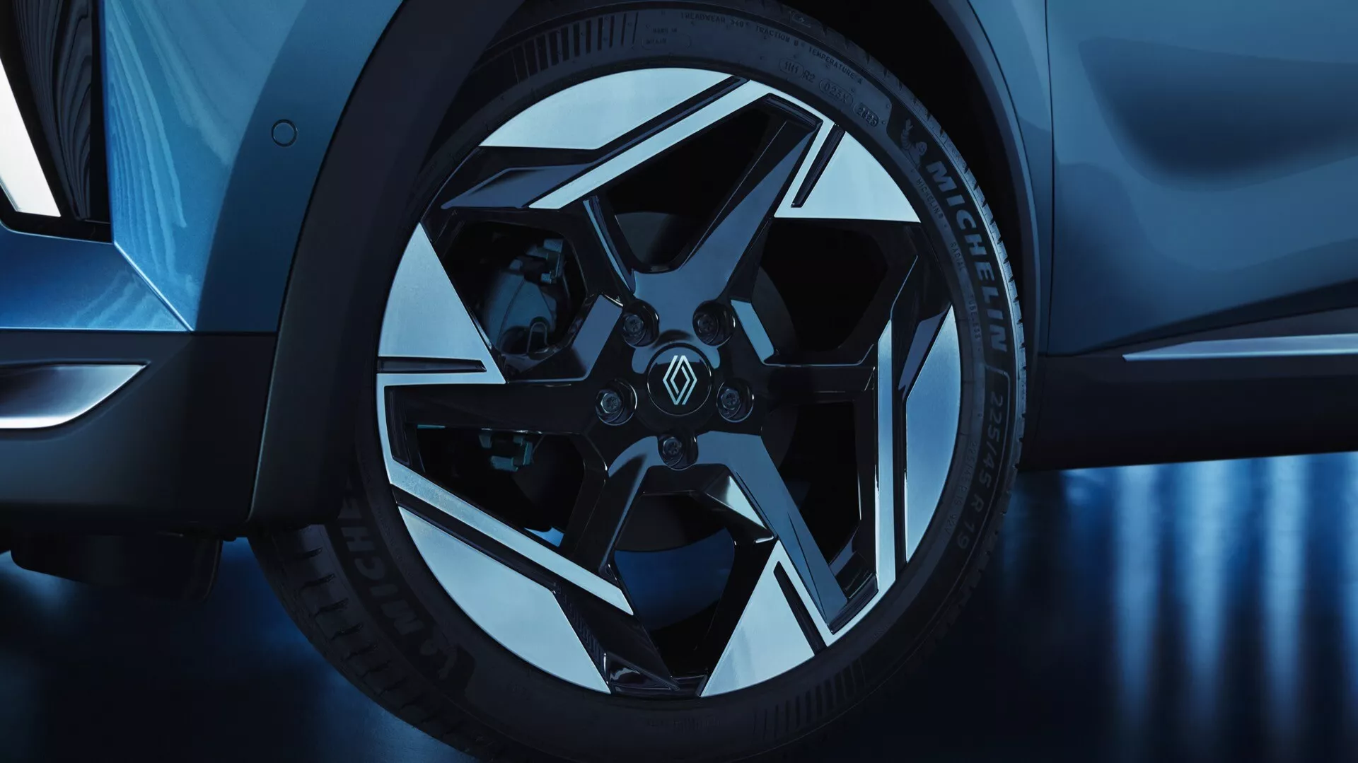 Detailný pohľad na predné koleso a čiastočný bočný profil Renault Symbioz 2024 (1. generácia) z nízkeho uhla. Koleso z ľahkých zliatin má zložitý, geometrický viacramenný dizajn s lesklým a matným povrchom, spojené s nízkoprofilovými vysokovýkonnými pneumatikami a značkovými strmeňmi, čo naznačuje športovosť a luxus. Chladný modrý tón zvyšuje estetiku, naznačuje štúdiové osvetlenie alebo postprocessing.