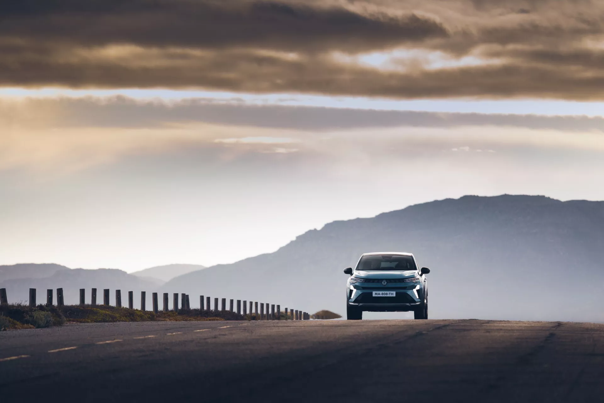 Pohľad z predného uhla na auto Renault Symbioz 2024 (1. generácia), zachytené počas skorých ranných hodín alebo neskorého popoludnia s teplým osvetlením a dlhými tieňmi. Vozidlo s elegantnými dizajnovými líniami a výraznými svetlometmi sa pohybuje smerom k fotoaparátu na vysoko položenom ceste, s mierne rozostrenými horami v pozadí. Osobitne osvetlené oblačné nebo dotvára atmosférický kontext; nízka perspektíva zdôrazňuje predné časti, ako je mriežka, kapota a nárazník. Pohybové rozmazanie na okrajoch ilustruje auto v pohybe, pričom zachováva ostré zameranie na modernú techniku a dizajn.