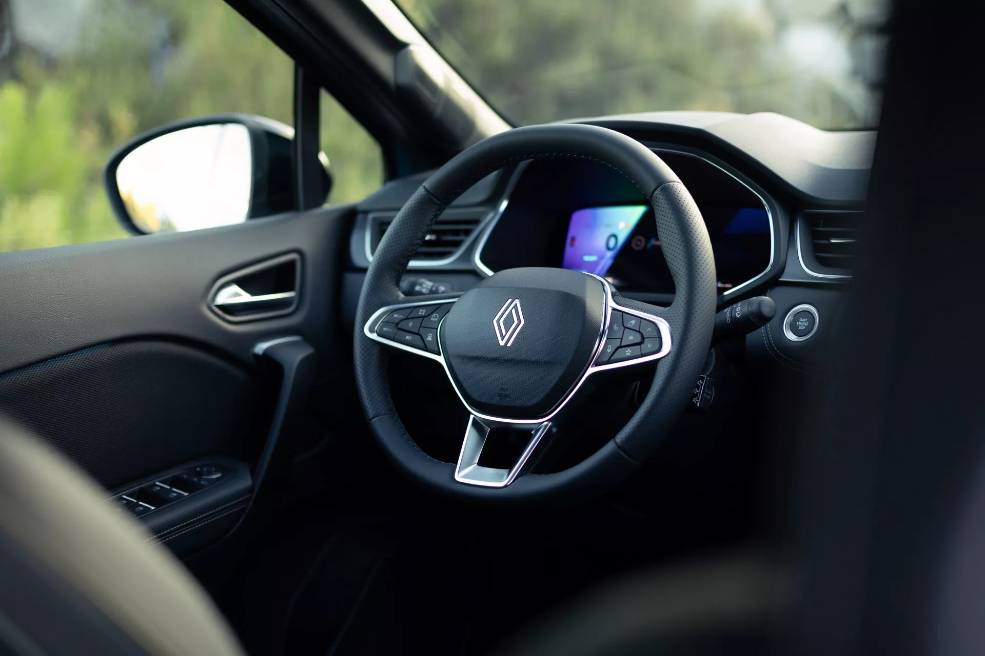 Pohľad do interiéru vozidla Renault Symbioz 2024 zo zadného sedadla, zameraný na moderný trojramenný volant a digitálnu prístrojovú dosku. Volant má ploché dno, kovový akcent a integrované ovládacie tlačidlá, všetko v tmavom farebnom schéme. Plytká hĺbka ostrosti zdôrazňuje tieto prvky, rozmazané okolie vrátane opierky hlavy a vonkajšej scenérie viditeľnej cez okno naznačuje, že vozidlo je v pokojovom stave.