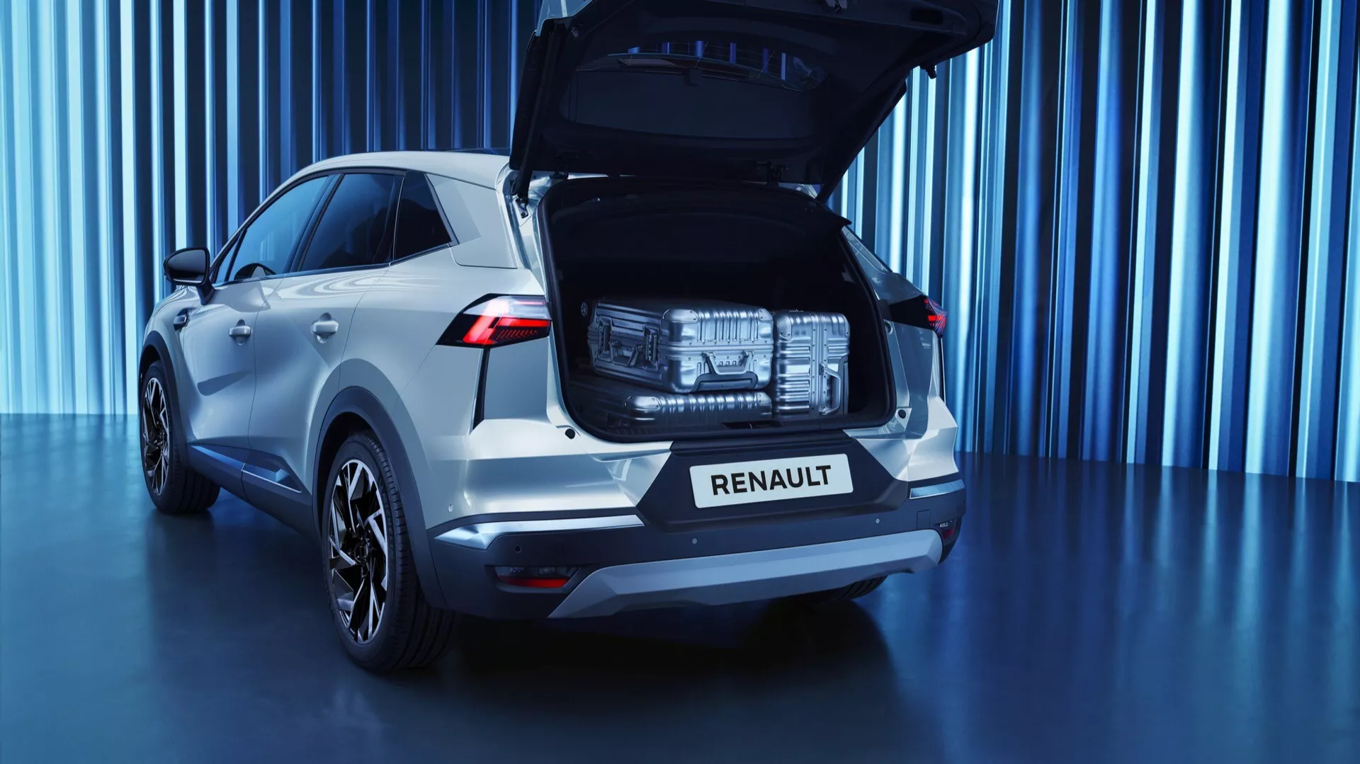 Zadný pohľad na strieborné SUV Renault Symbioz 2024 zaparkované pred modrým pozadím s vertikálnym svetelným pruhom. Auto, fotografované z nízkeho uhla, zdôrazňuje veľkosť a dizajn zadného nárazníka, zadných svetiel a celkovú zadnú estetiku. Otvorený zadný výklopný kryt odkrýva nákladný priestor, čo poukazuje na skladovacie schopnosti. Obrázok obsahuje viditeľné moderné LED zadné svetlá a integrované parkovacie senzory v zadnom nárazníku, čo naznačuje pokročilé systémy asistencie vodičovi. Viacramenné zliatinové disky naznačujú vyššiu úroveň výbavy alebo športový balík. Umelecký nízkoúhlový záber zachytáva prvky dizajnu vozidla a technologické funkcie.