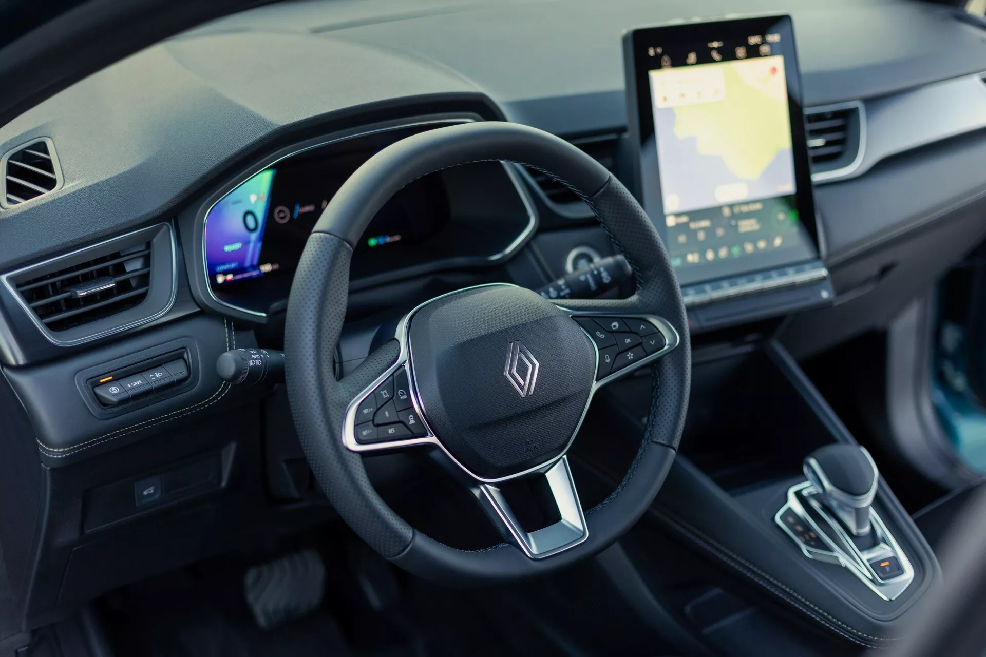 Záber interiéru Renault Symbioz 2024 (1. generácia) z pohľadu vodiča, zameraný na volant a palubnú dosku. Obrázok poskytuje pohľad z vtáčej perspektívy, 30 stupňov nadol, a zobrazuje trojramenný volant s multifunkčnými tlačidlami a obecným emblémom, plne digitálny prístrojový panel a veľký dotykový infotainmentový systém so zobrazením mapy. Stredová konzola zahŕňa moderný volič prevodovky s minimalistickým dizajnom. Interiér predstavuje vysoko technologický dizajn elektrického vozidla s ambientným osvetlením a modernou estetikou.