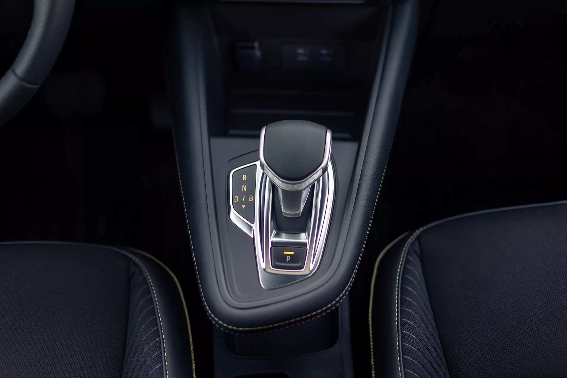 Pohľad zhora na stredovú konzolu Renault Symbioz 2024 so zameraním na elektronický volič prevodovky s chrómovým povrchom a osvetlenými indikátormi prevodov pre parkovanie, spiatočku, neutrál a jazdu. Volič má kožené boky, žlto zvýraznené tlačidlo parkovania a je umiestnený v lesklom čiernom puzdre s prepracovaným prešívaním okolitého koženého interiéru.