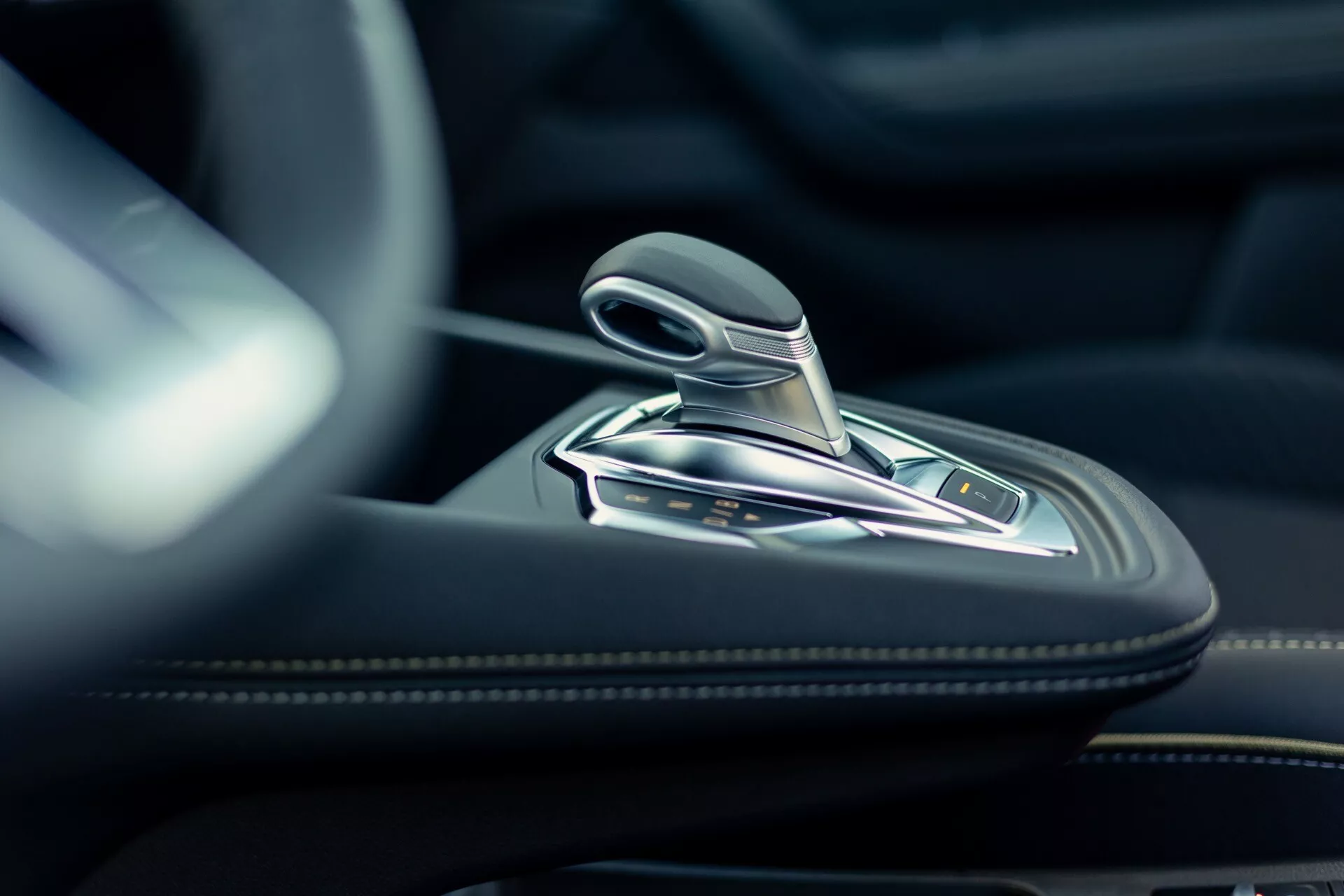 Detailný záber na vnútornú časť Renault Symbioz 2024 zobrazujúci modernú radiacu páku s kovovými prvkami a pravdepodobne koženým povrchom. Vysokorozlíšený obrázok urobený z nízkeho uhla sa sústredi na automatickú radiacu páku a okolitú stredovú konzolu vyrobenú z vysoko kvalitných materiálov, ako je koža, brúsený kov alebo chróm. Na konzole sú viditeľné rôzne tlačidlá pravdepodobne pre jazdné režimy, parkovaciu brzdu, funkciu auto-hold a kontrolu trakcie. Mäkké, prirodzené svetlo zvýrazňuje luxus a presné inžinierstvo interiéru auta.