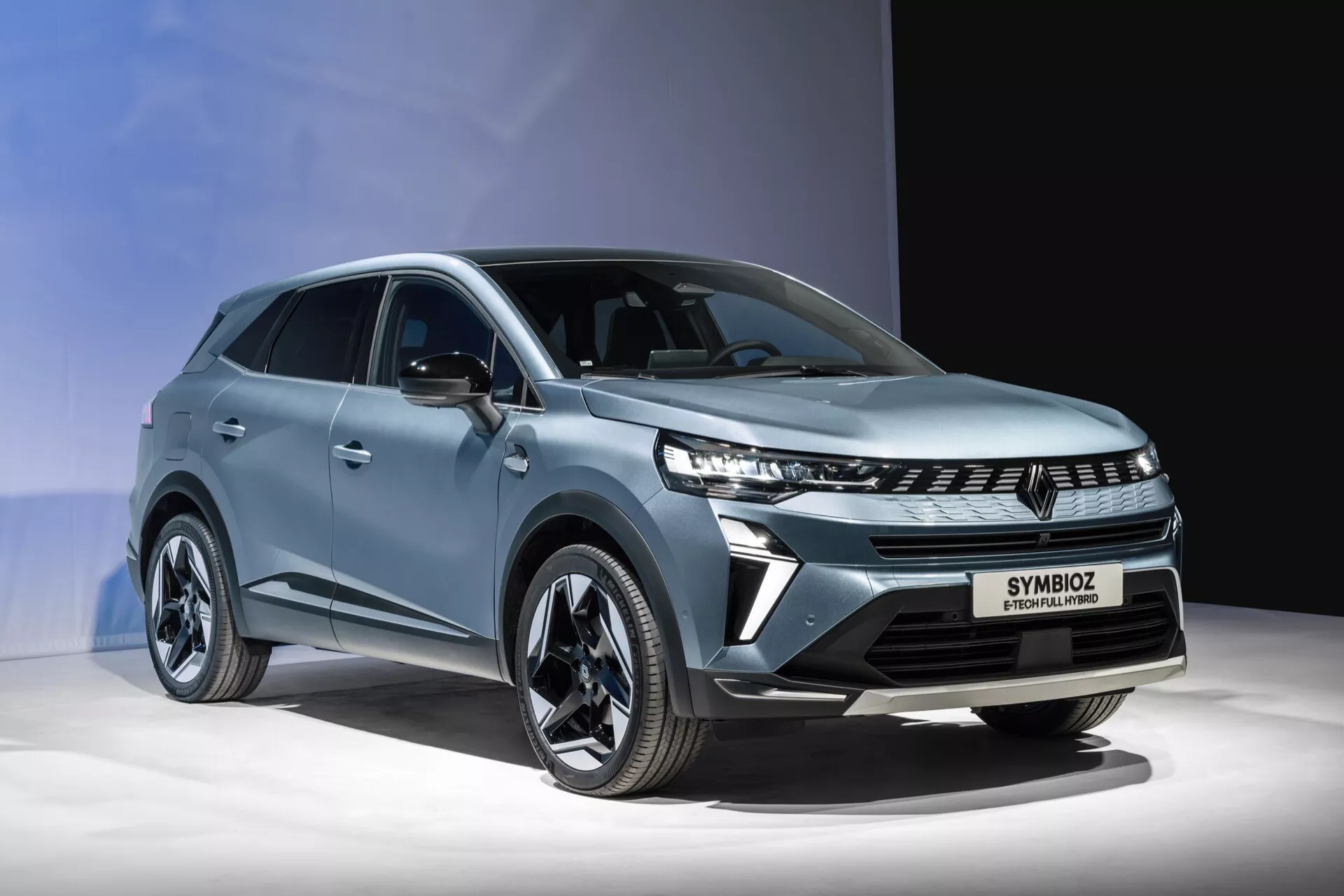 Tento obraz zachytáva Renault SYMBIOZ 2024, prvej generácie, predstavený v modernom SUV dizajne na dobre osvetlenom pódiu, pravdepodobne na autosalóne alebo propagačnej akcii. Predný uhol tri štvrtiny odhaľuje jeho štýlové LED svetlomety, charakteristickú osvetlenú mriežku, veľké zliatinové kolesá so súčasným dizajnom lúčov a ostré geometrické línie karosérie. Svetlomodrý kovový povrch dopĺňa aerodynamický obrys vozidla, s jasne viditeľným označením 'SYMBIOZ', ktoré naznačuje pokročilú technológiu pohonu, pravdepodobne elektrickú alebo hybridnú. Zatemnené okná a integrované smerovky v bočných zrkadlách ešte viac zdôrazňujú jeho prémiový a sofistikovaný charakter.