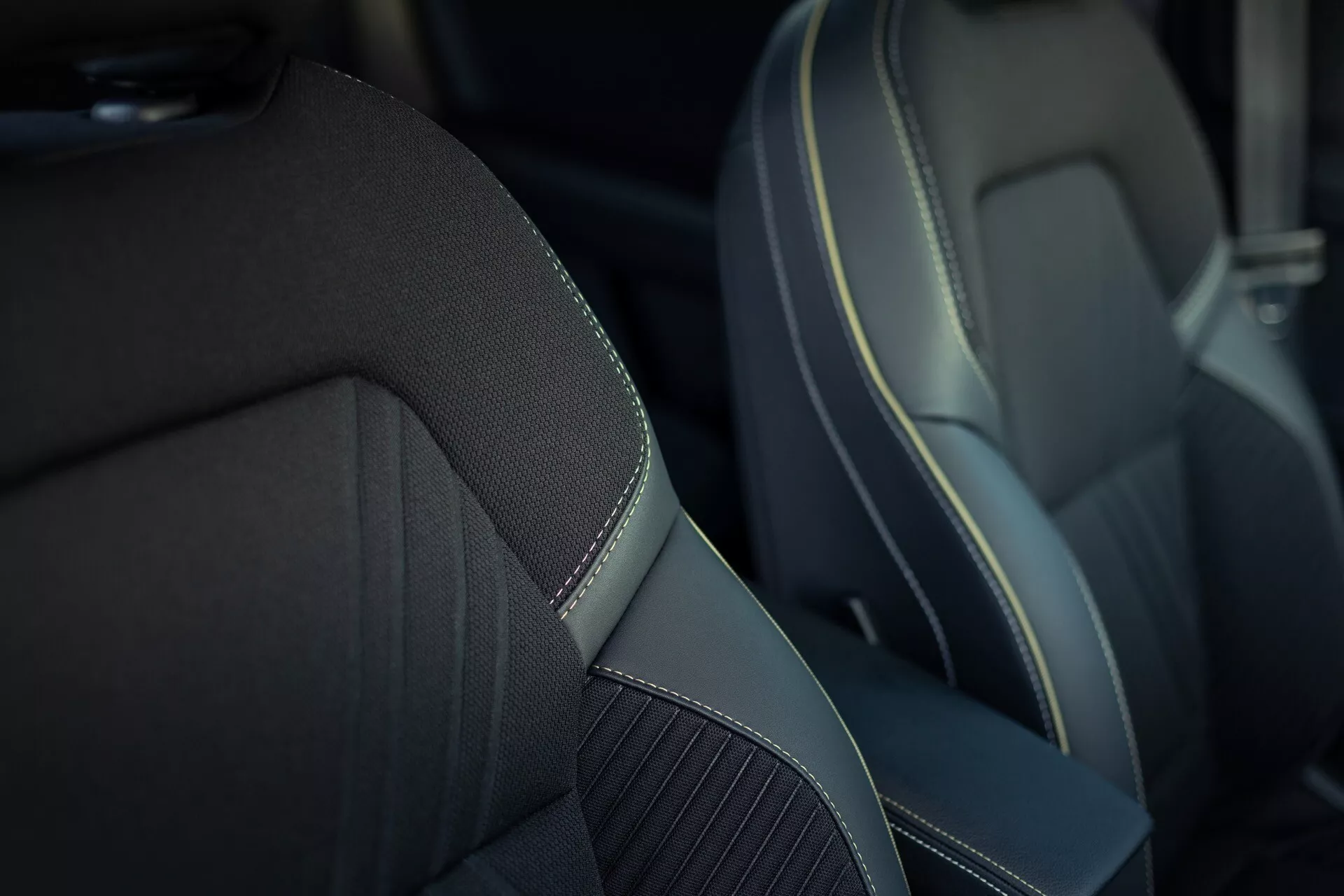 Blízky pohľad na predné sedadlo spolujazdca v Renault Symbioz 2024 (1. generácia). Sedadlo má stredový panel vyrobený z textúrovanej látky alebo syntetického tkaniva pre lepšiu priedušnosť, obklopený hladkým syntetickým materiálom podobným koži s kontrastným ozdobným šitím, ktoré zvýrazňuje prémiový alebo športový dizajn. Plytká hĺbka ostrosti sa zameriava na sedadlo a rozmazáva pozadie, aby vynikla kvalita materiálu a remeselné spracovanie.