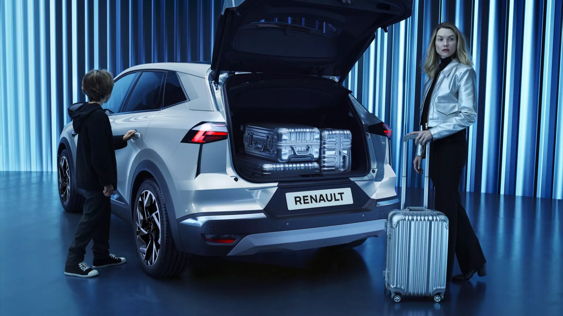 Trojštvrťový zadný uhol pohľadu na biely SUV Renault Symbioz 2024 v showroome, s otvoreným zadným vekom ukazujúcim priestranný kufor, moderné LED zadné svetlá, logo Renault pod zadným čelným sklom, parkovacie senzory na nárazníku, kolesá zliatin s viacerými lúčmi, systém automatického otvárania na tlačidlo a dvomi osobami: žena s kufrom, naznačujúcou cestovanie a dieťa, ktoré interaguje s kufrom, zdôrazňujúcim rodinné aspekty vozidla.