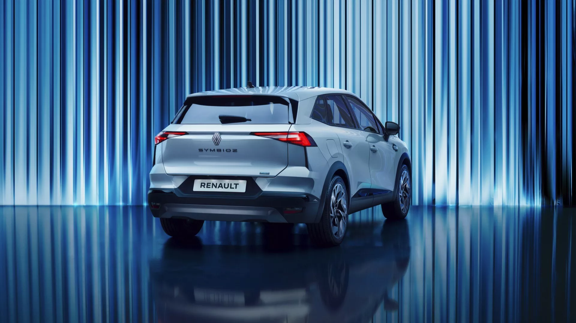 Tento profesionálny snímok zobrazuje zadný koniec prvej generácie Renault Symbioz 2024, moderného SUV modelu. Vozidlo má charakteristické LED zadné svetlá, zadný spojler, zadný stierač, znak Renault a názov modelu. Záber z nízkeho uhla zdôrazňuje jeho postoj a zvýrazňuje čistý dizajn bez viditeľného výfuku, čo naznačuje hybridný alebo elektrický pohon. Na živom pozadí s vertikálnymi modrými svetelnými pruhmi, svetlý metalický odtieň vozidla a reflexná podlaha vytvárajú sofistikovanú atmosféru showroomu alebo inscenovaného fotenia.