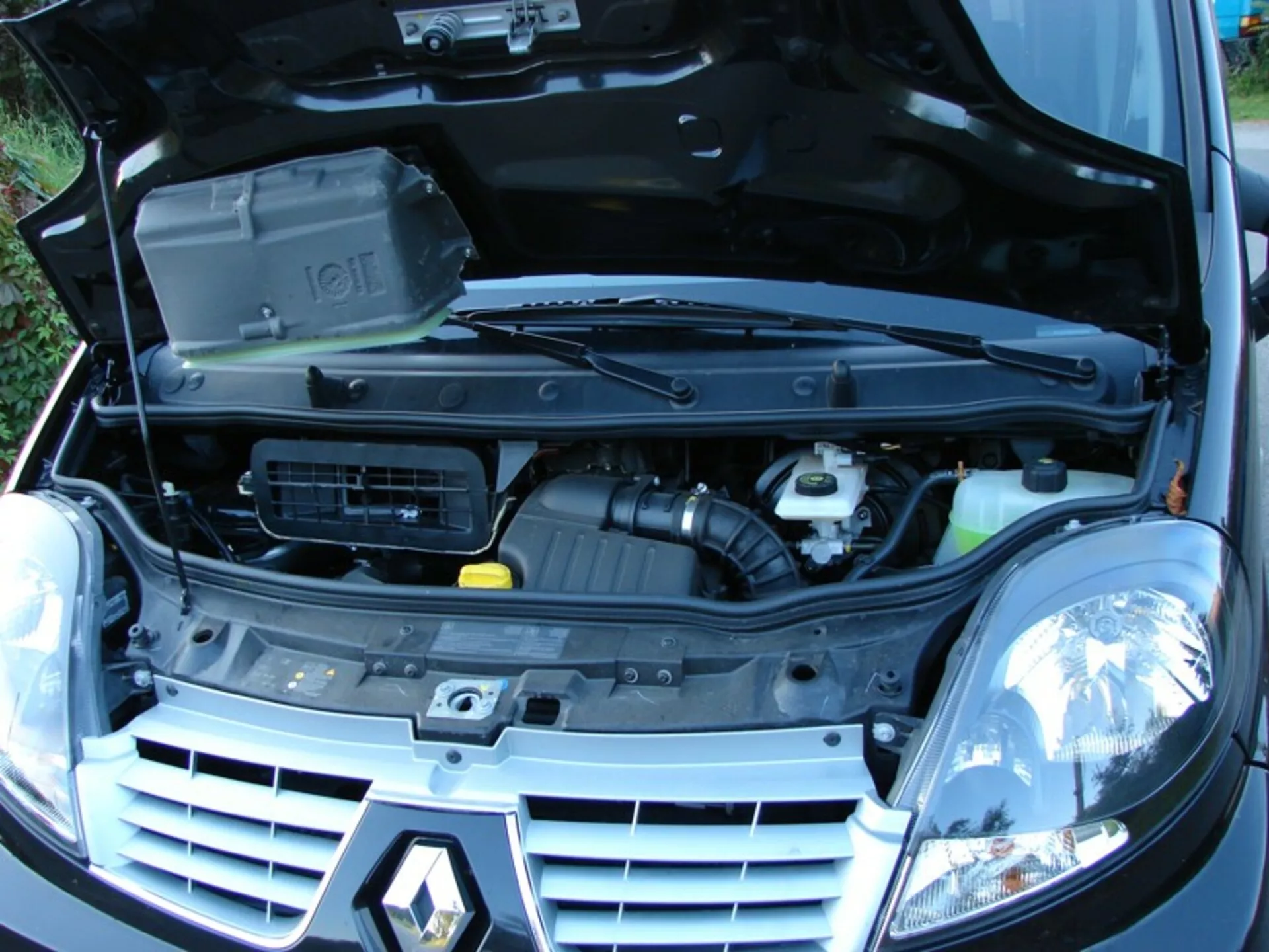 Predný trojštvrťový pohľad na Renault Trafic Combi z roku 2006 s otvorenou kapotou odhaľujúci motorový priestor a komponenty, vrátane bloku motora s krytom, prívodu vzduchu a nádrží na kvapaliny, obklopený tradičnými halogénovými svetlometmi