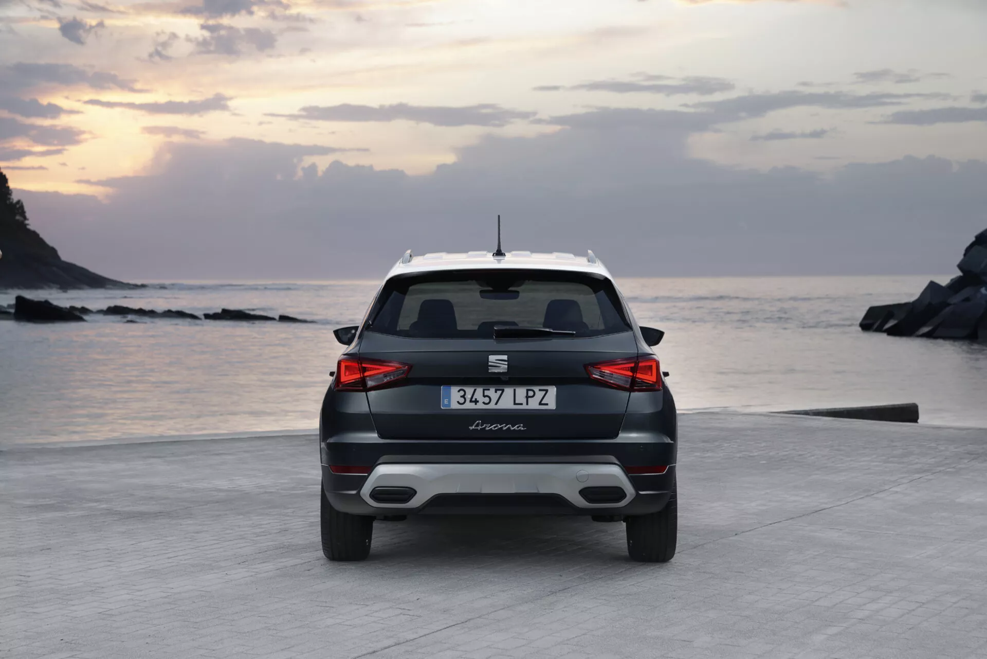 Zadný pohľad na moderný Seat Arona 2017 za úsvitu alebo súmraku s mäkkým osvetlením, športovým zadným spojlerom, elegantnými vodorovnými zadnými svetlami a možnými parkovacími senzormi na pobrežnej ceste blízko pláže a skalnatého pobrežia.