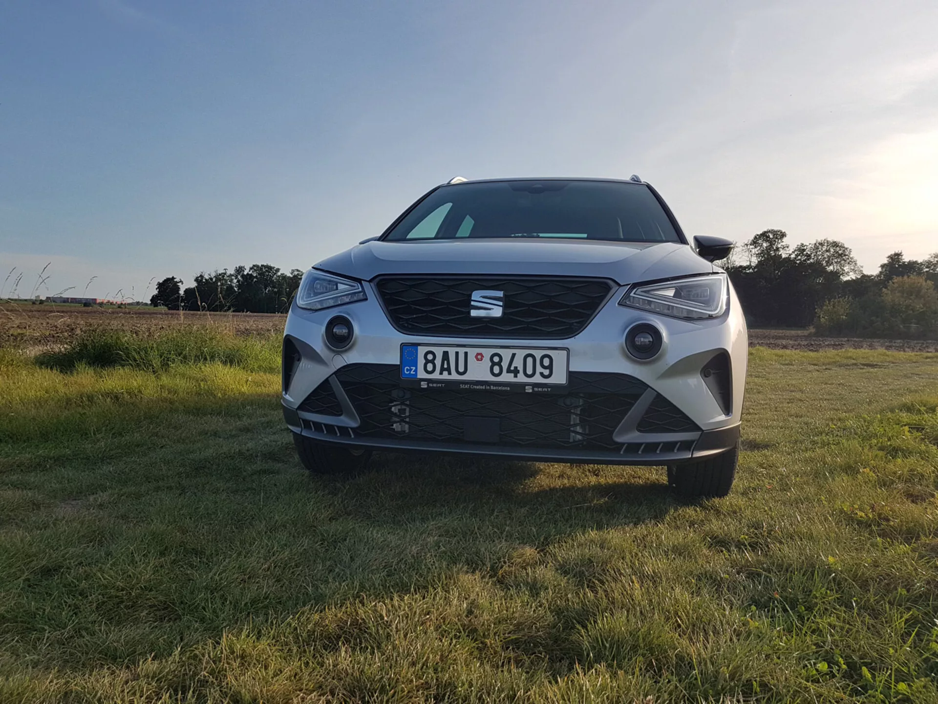 Seat Arona 2017 fotografovaný z predného uhla na úrovni terénu, ukazujúci jeho LED svetlomety, výraznú prednú mriežku so znakom, veľký vzduchový vstup, parkovacie senzory na nárazníku a hmlové svetlá v uhlových obaloch proti pozadiu trávnatého poľa osvetleného západom slnka