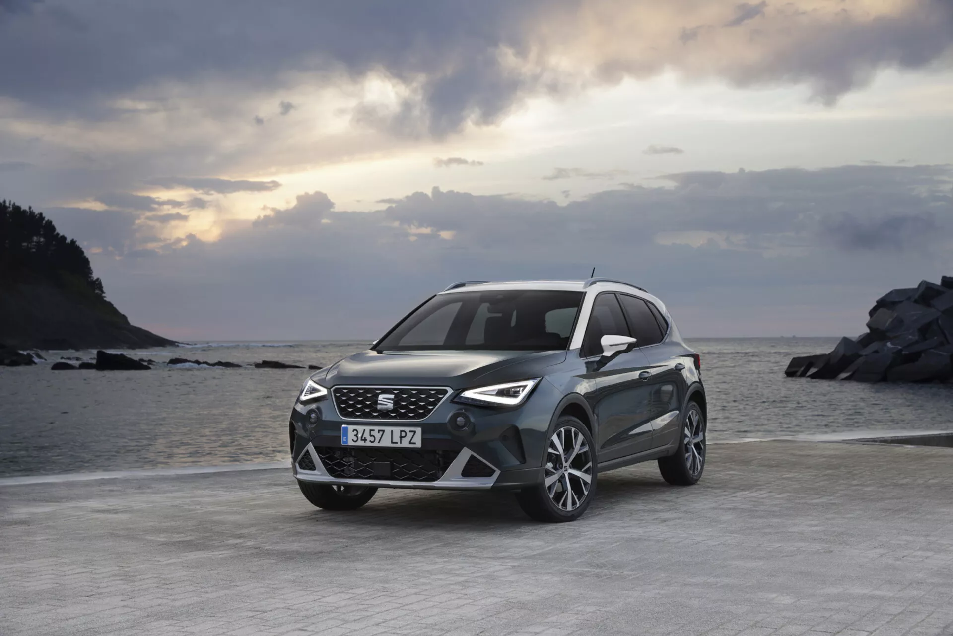 Crossover/SUV Seat Arona z roku 2017 zaparkovaný na rovnatom teréne s výhľadom na morskú a skalnatú krajinu na pozadí počas svitania alebo súmraku s čiastočne zamračeným nebom, predstavujúci jeho pohľad z prednej trojštvrtiny, čo zdôrazňuje moderný dizajn mriežky a LED svetlometov, športový predný nárazník, zliatinové kolesá a metalický lak.