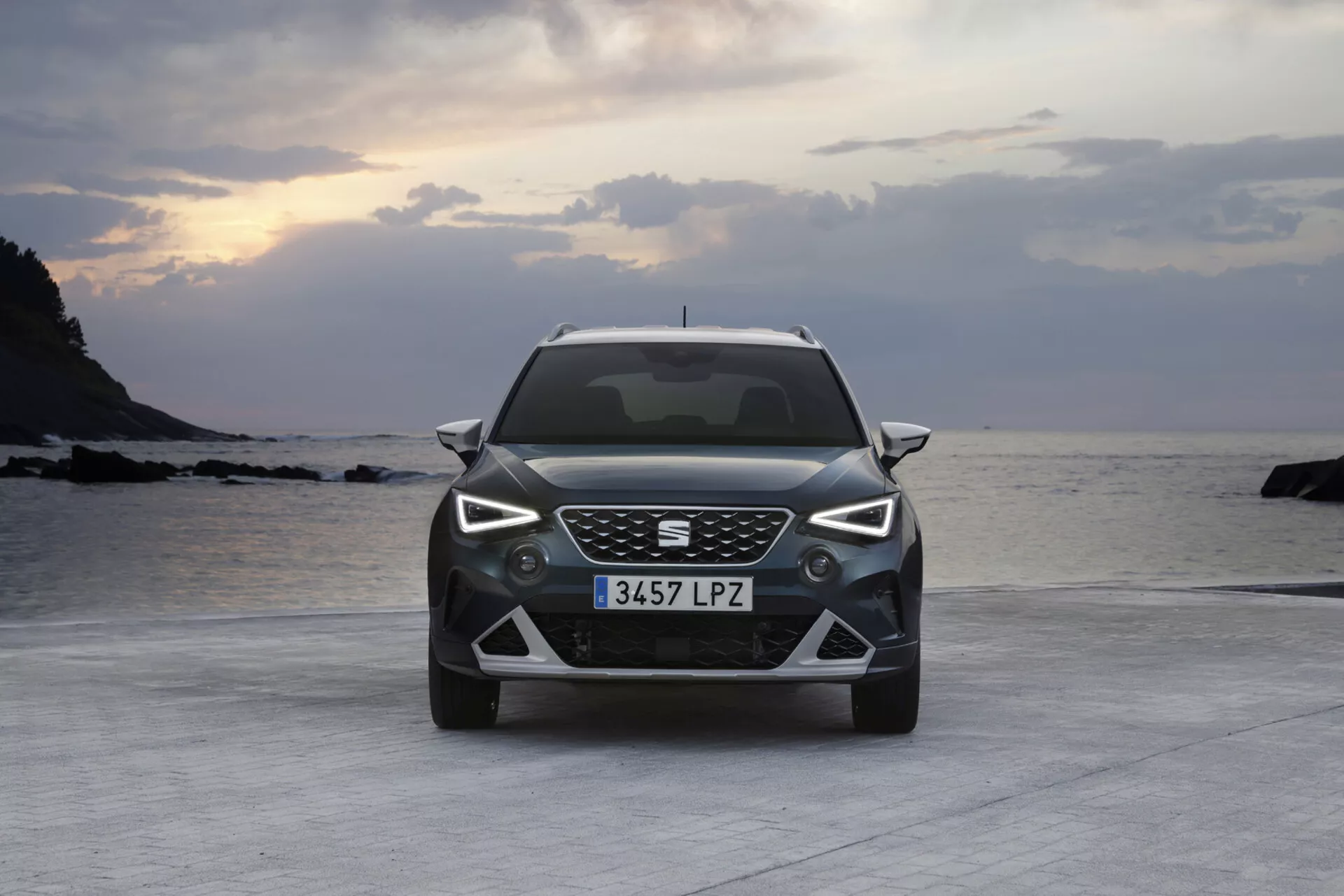 Seat Arona 2017 zobrazené z predného uhla, ktoré ukazuje jeho modernú mriežku s včelím vzorom, LED svetlomety, detailný spodný nárazník a zliatinové disky, zaparkované na spevnenej ploche blízko pláže s oceánom a súmrakovou oblohou na pozadí.