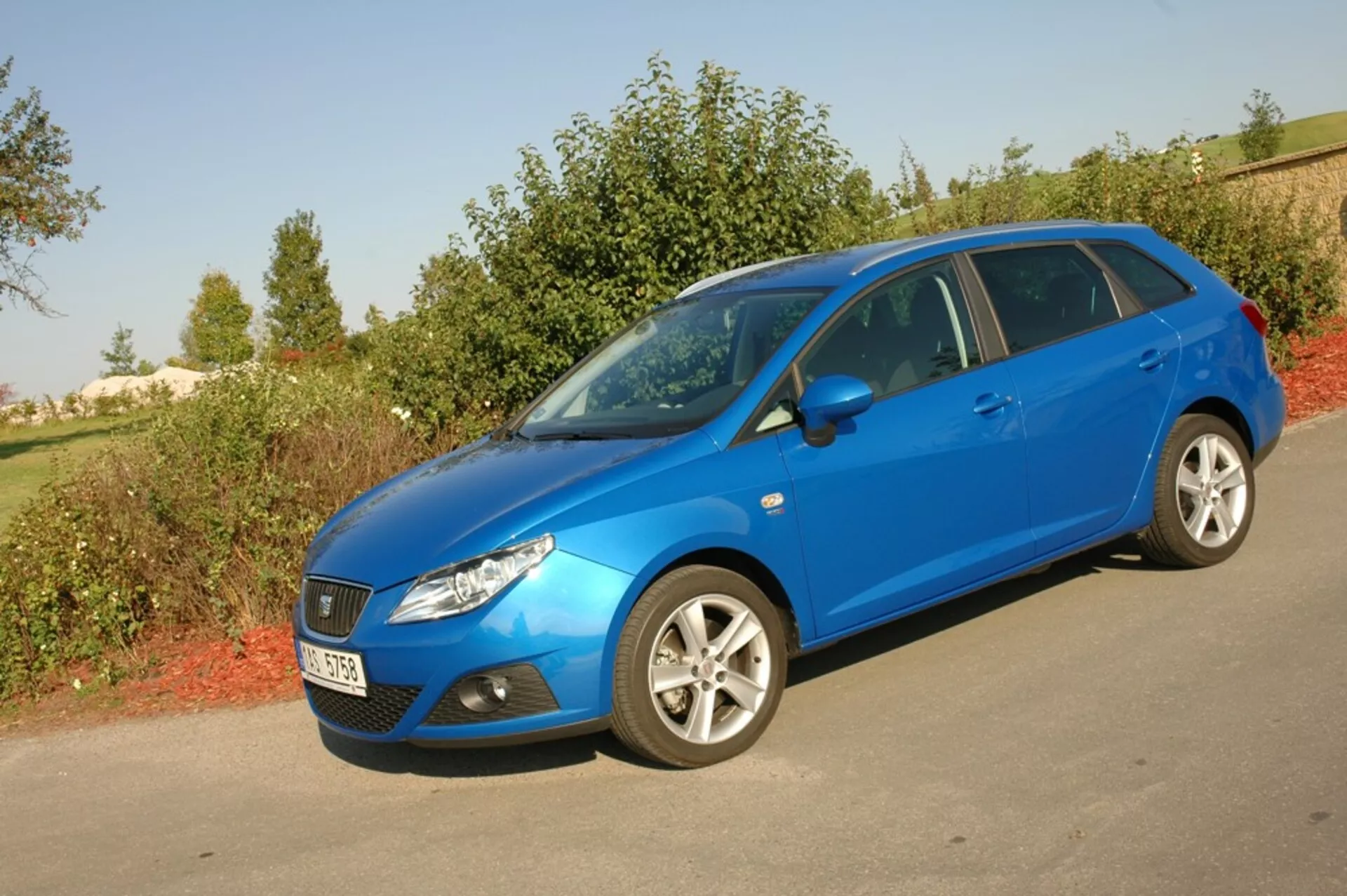 Svetlomodrý hatchback Seat Ibiza 2012 zaparkovaný na asfalte oproti zeleni, predvádzajúci svoju aerodynamickú prednú mriežku, svetlomety natiahnuté dozadu a bočný profil s viacprvkovými zliatinovými kolesami pod prirodzeným denným svetlom