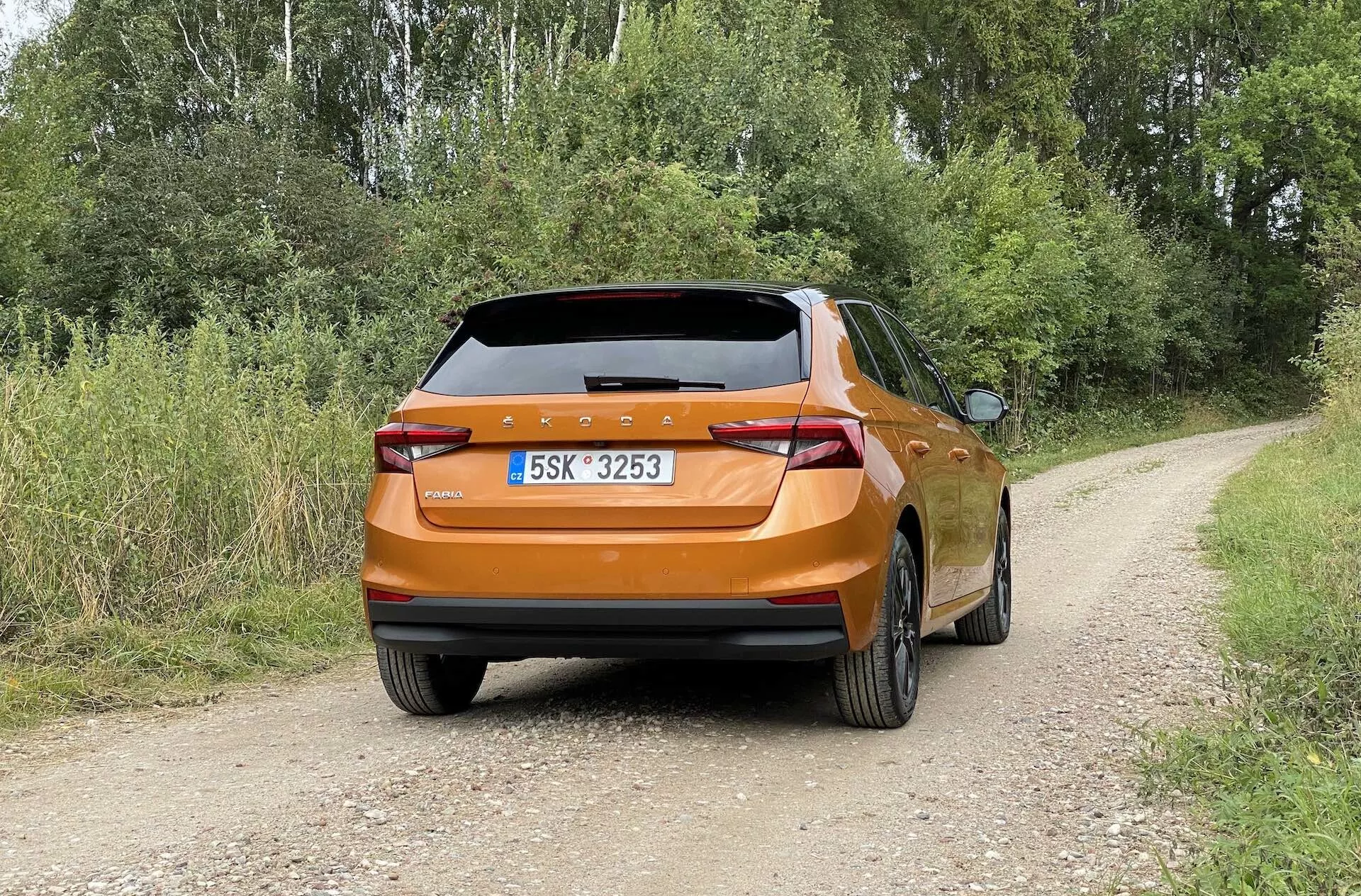 Zadný pohľad na oranžovú Škoda Fabia 2021 na nespevnenej vidieckej ceste obklopenej listami a stromami. Obrázok ukazuje auto za denných svetelných podmienok s oblačnou oblohou, zvýrazňujúc moderné LED zadné svetlá, stredovú poznávaciu značku, dvojité výfukové otvory a zadné sklo s stieračom. Vozidlo je umiestnené v strede a je nehybné s pevným dotykom pneumatík na zemi.