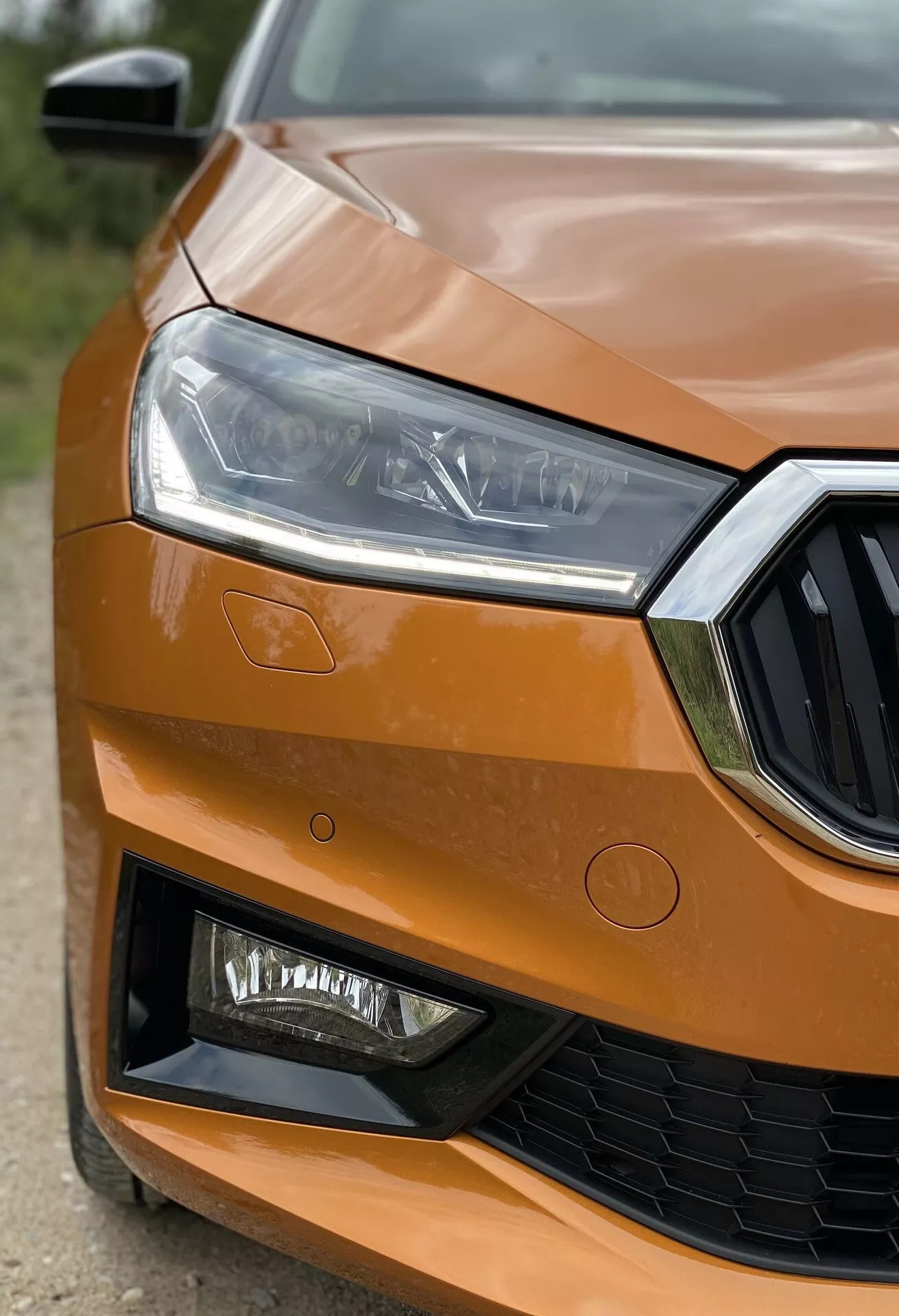 Detailný záber na prednú ľavú časť Škody Fabia 2021, zvýrazňujúci agresívny a moderný dizajn z predného štvrtinového uhla. Elegantný dizajn LED svetlometov, čiastočne viditeľná mriežka s úchytom centra znaku, metalíza oranžová farba zdôrazňujúca luxus a ostré úhly nárazníka s otvormi a potenciálnymi hmlovými svetlami, ktoré zvýrazňujú technické a výkonnostné vlastnosti vozidla.