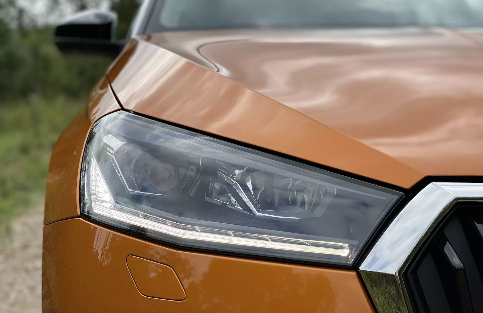 Detailný záber na predný ľavý roh Škody Fabia 2021, zvýrazňujúci elegantný dizajn svetlometov s viacerými svetelnými prvkami a časť veľkej, hranatej mriežky. Predná časť je zachytená z čelného šikmého uhla, ukazujúca časť metalickej oranžovej kapoty s dynamickými kontúrami a hladkými vonkajšími panelami. Fotografia zdôrazňuje svetlomety a mriežku s ostrým popredím a jemne rozmazaným pozadím.