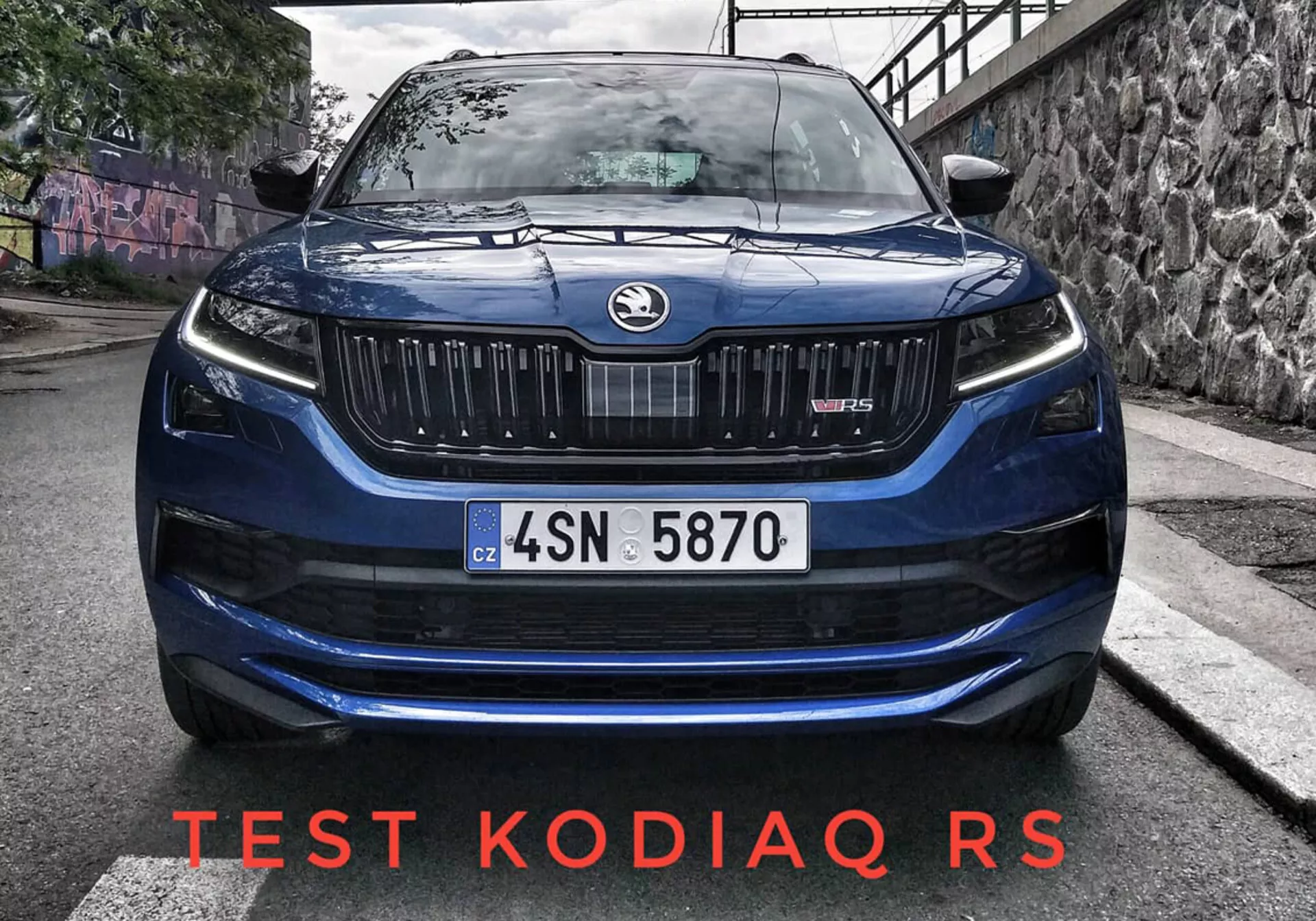 Zblízka pohľad zpredu na Škoda Kodiaq Scout 2017 s dominantnou mriežkou s vertikálnymi lamelami, elegantnými LED svetlometmi, jemne vyklenutou kapotou pre zlepšenie aerodynamiky a efektívnymi sacími otvormi na nárazníku na mestskom ceste s graffiti na pozadí a prirodzeným denným svetlom