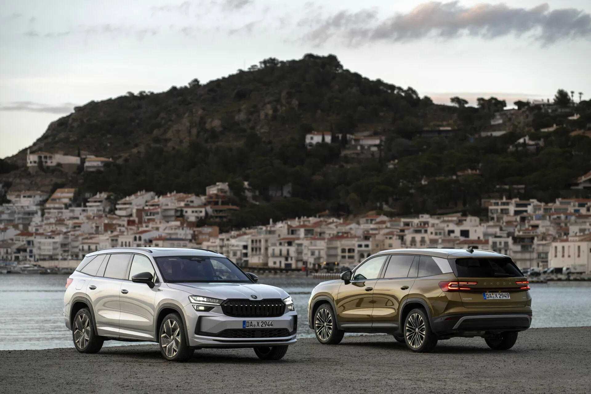 SUV Skoda Kodiaq 2024 zaparkované vedľa seba na nábrežnej promenáde pri svitaní s pozadím pobrežného mesta, predvádzajúce prednú mriežku, svetlomety a štýlové dizajnové línie jedného a zadný trojštvrťový pohľad na druhé.