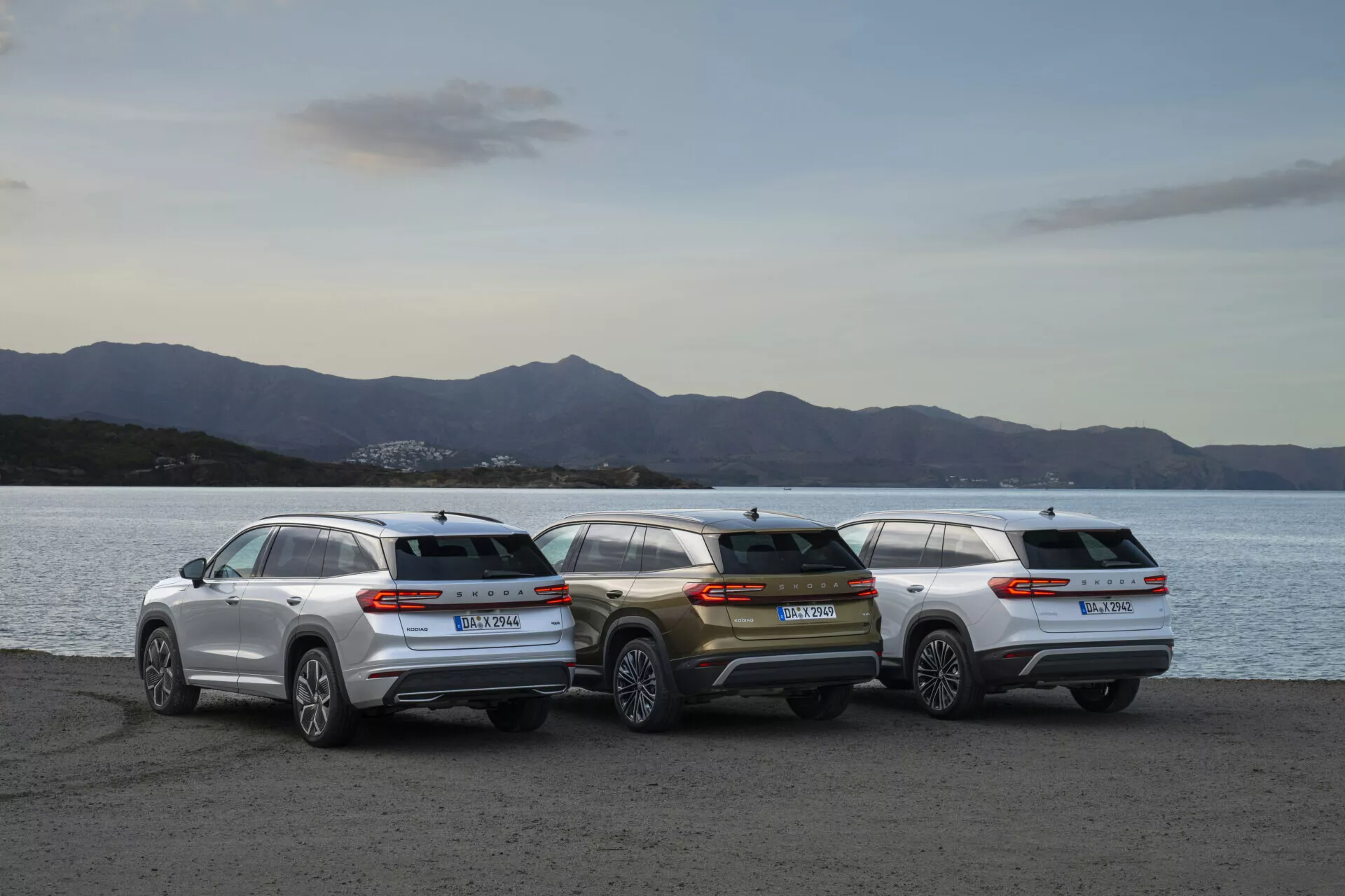 Rad Skoda Kodiaq 2024 s tromi SUV parkovanými na nábreží počas súmraku, predvádzajúce zadné profily a dizajnovú estetiku na pozadí jemne osvetleného pozadia
