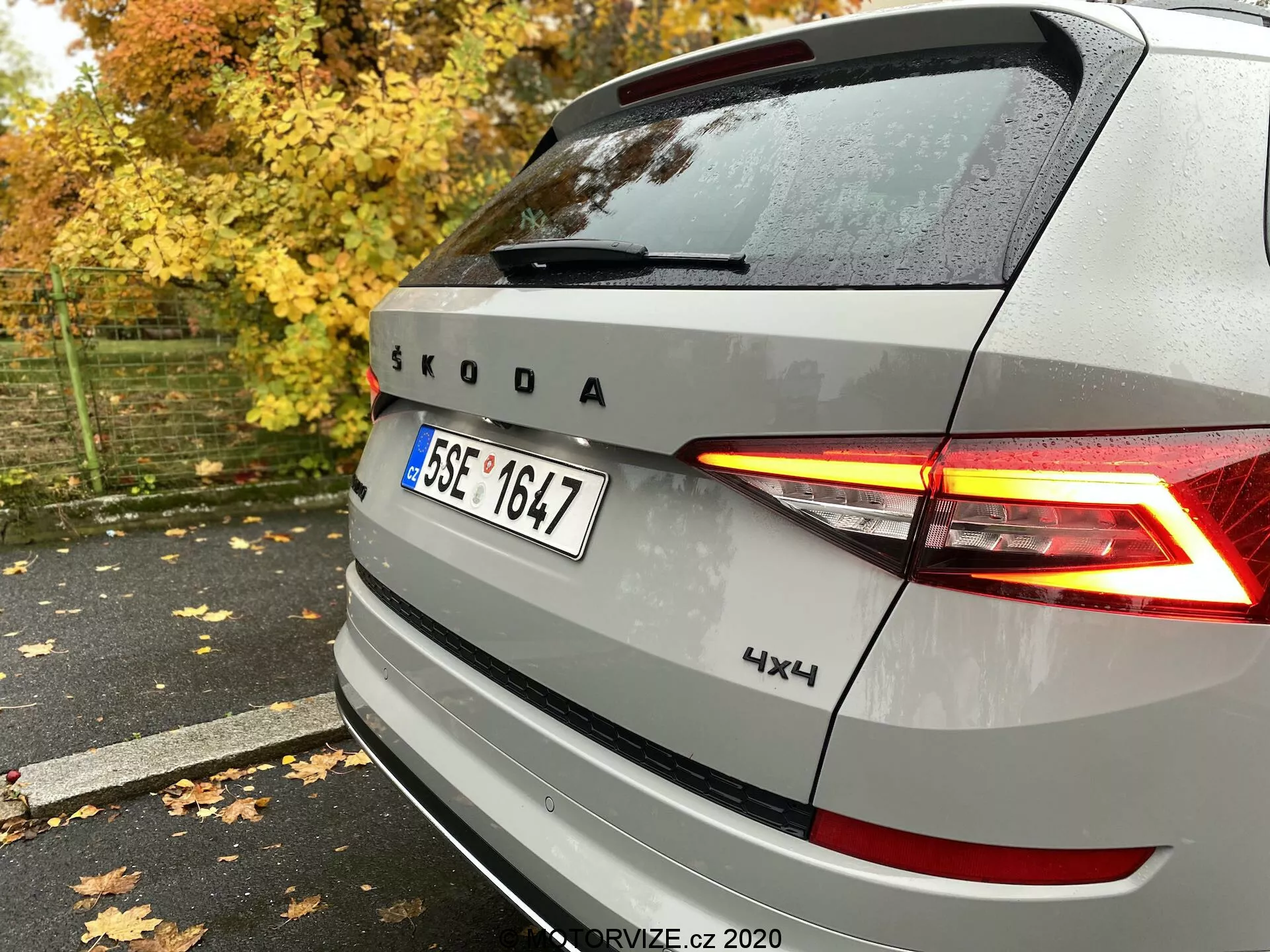 Zadný pohľad na strieborné vozidlo Škoda s osvetlenými zadnými LED svetlami v tvare C, znakom Škoda a značkou, s označením 4x4, zachytený zozadu sprava na mokrom chodníku medzi jesennými listami a stromami.