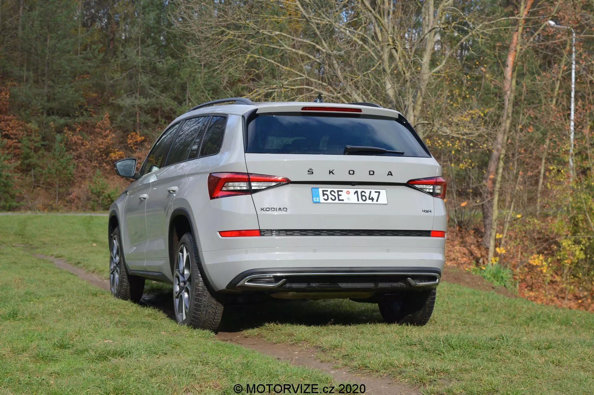 Pohľad na strieborný Škoda Kodiaq SUV z roku 2017 zo zadného trojštvrťového uhla, zaparkovaný na trávnatej ploche s pozadím stromov. Vozidlo má strešný spojler, žraločiu anténu, zadný stierač a odmrazovače okien. Na zadných dverách je viditeľné logo Škoda a modelový názov 'Kodiaq', s európskou poznávacou značkou. Obvinovacie zadné svetlá naznačujú LED technológiu. Nárazník obsahuje parkovacie senzory, odrazky a zadný kryt. Viditeľné sú obrysy dvojitého výfuku a bočný profil ukazuje zliatinové kolesá a ochranné lišty.